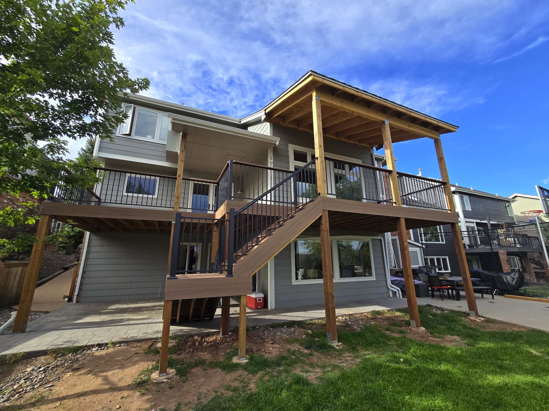 A multi-story house with a newly built wooden deck and staircase, black metal railings, a sliding glass door, and outdoor furniture. The yard has grass and dirt patches, with other houses visible in the background under a blue sky with clouds.