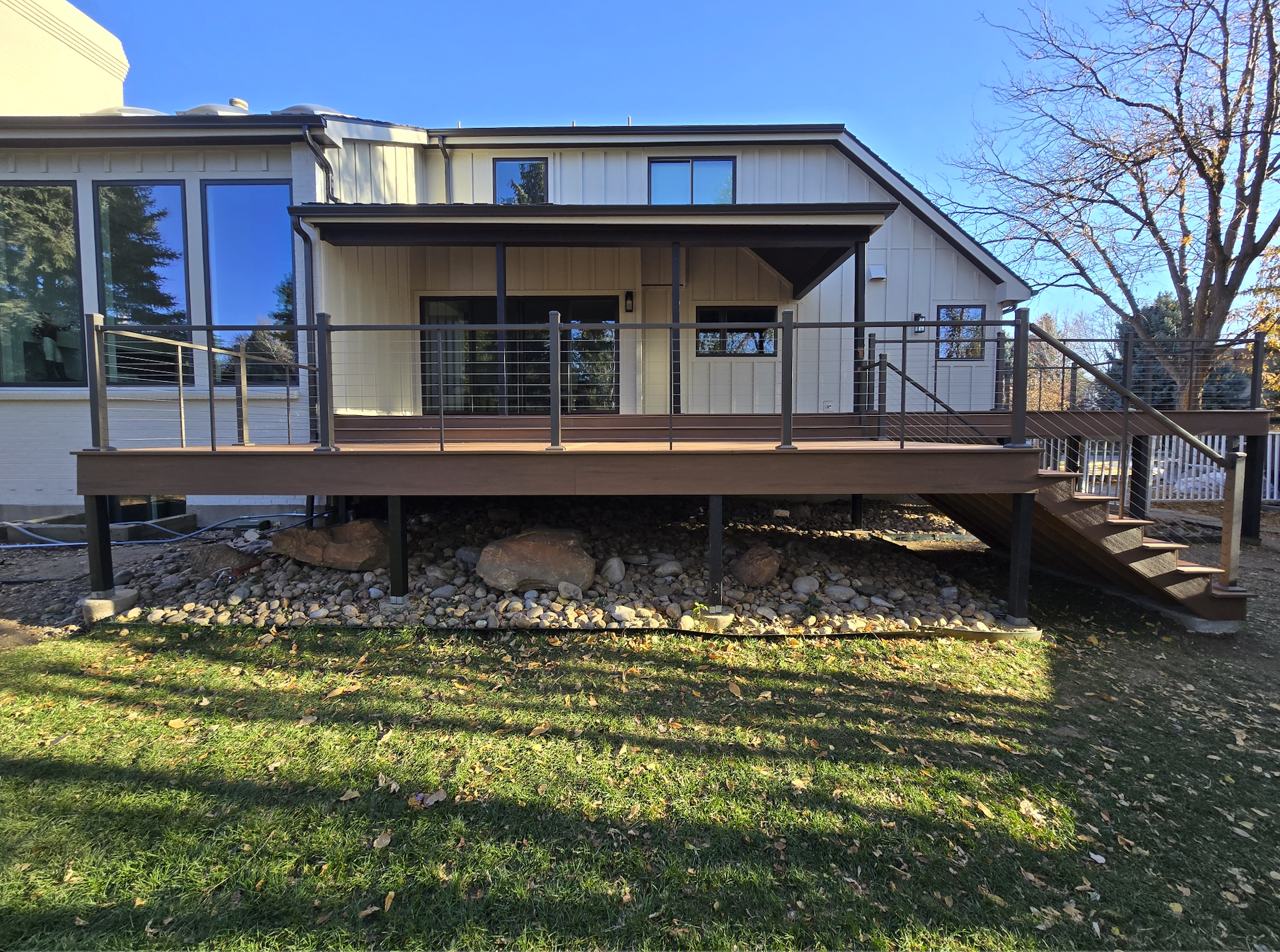 A modern two-story house with white siding and large windows, featuring a spacious wooden deck with metal railings, and outdoor stairs leading to a grassy yard with rocks underneath.