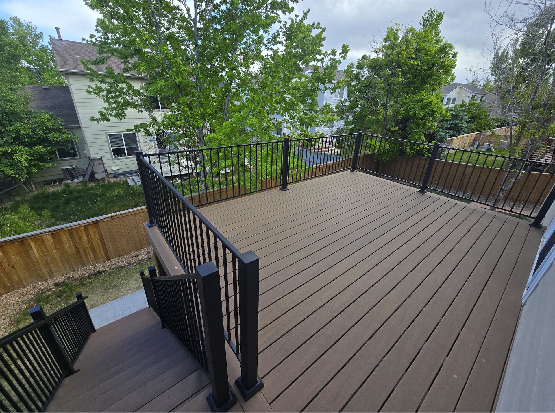 Empty wooden deck with black metal railing, elevated above yard with trees and neighboring houses in the background. Trex decking. Highlands Ranch 