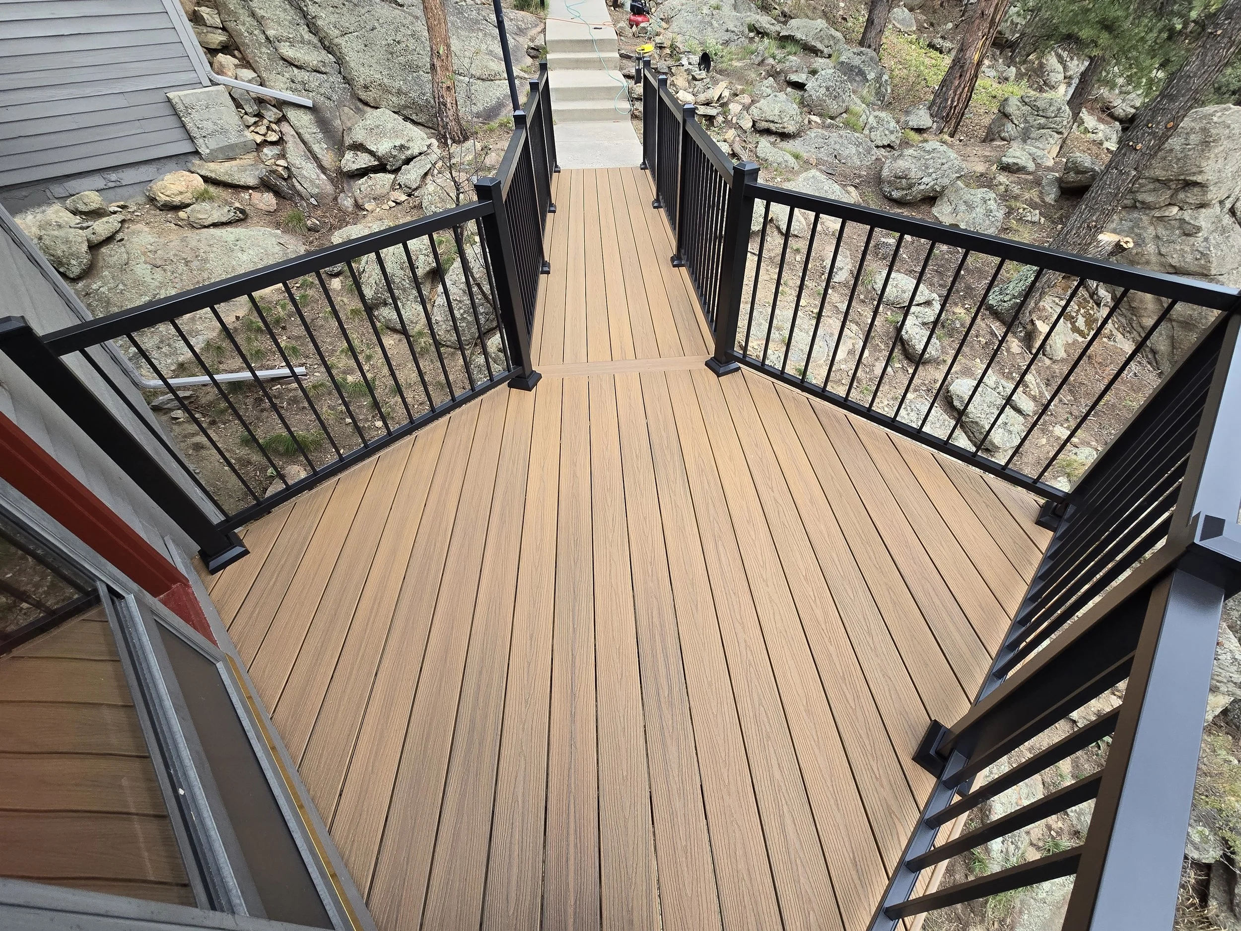 A wooden deck with black metal railings surrounding it, built on a slope with large rocks and trees in the background.
