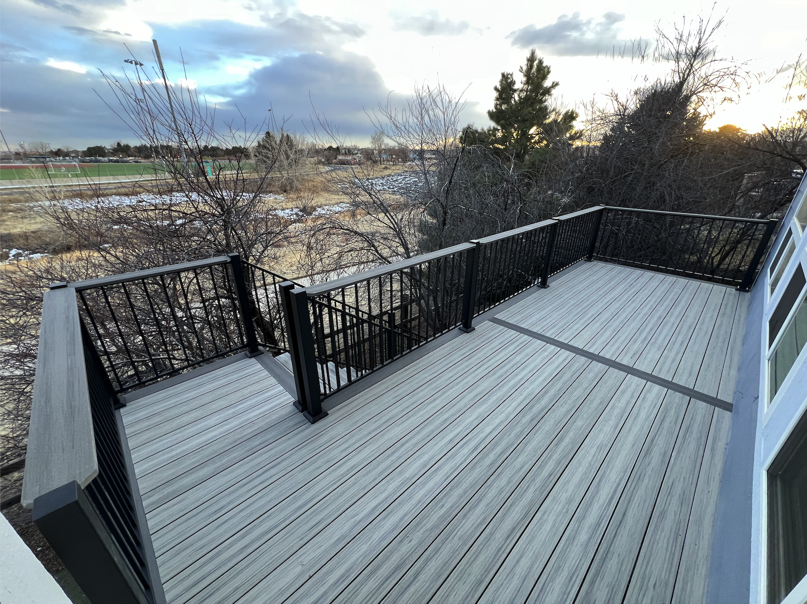 View of a modern deck with light gray Deckorators decking and black metal railing, overlooking trees and a park with a sports field in the distance during sunset, with some snow on the ground. Parker 