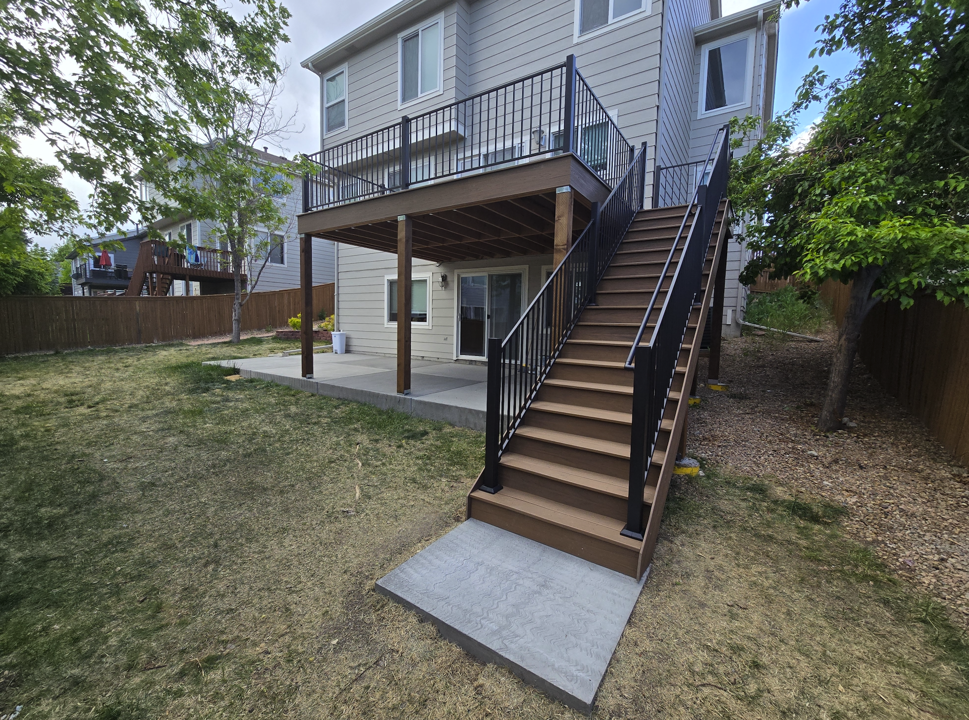 Backyard with a two-story house featuring a wooden deck and staircase, surrounded by a wooden fence, trees, and a grassy yard. Trex decking. Highlands Ranch