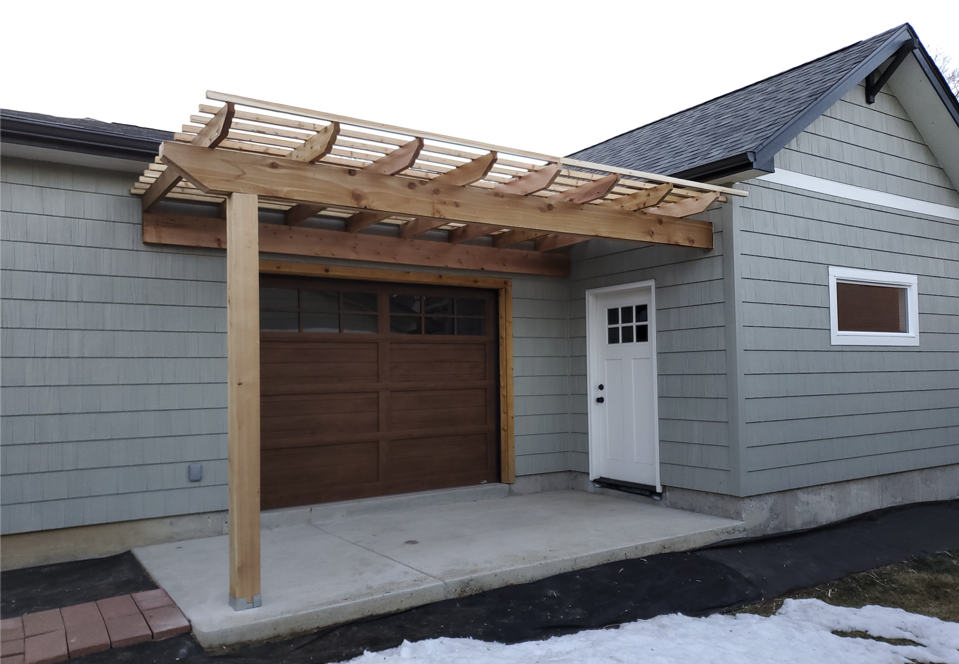 Carriage house with a new wooden pergola under construction, white door, window, concrete patio, and snow on the ground.