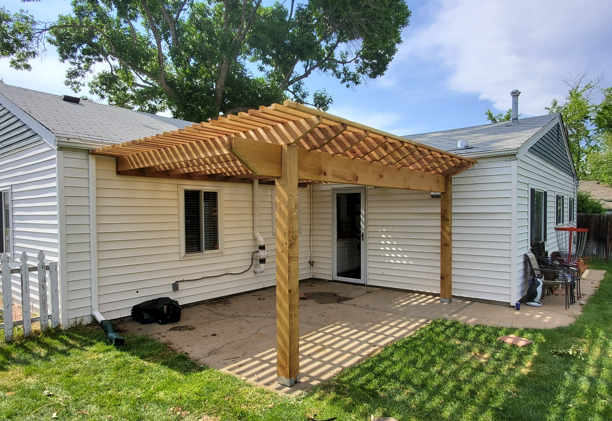 A white house with vinyl siding and a gray shingled roof. There is a newly built wooden pergola with a slatted roof over a concrete patio. The yard has green grass and there are some outdoor chairs and a basketball hoop in the background.