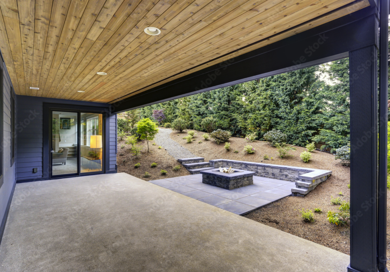View of a backyard patio with a fire pit, stone steps, and a landscaped hill with trees and shrubs, seen from under a covered patio with wooden ceiling, adjacent to a house with sliding glass doors.