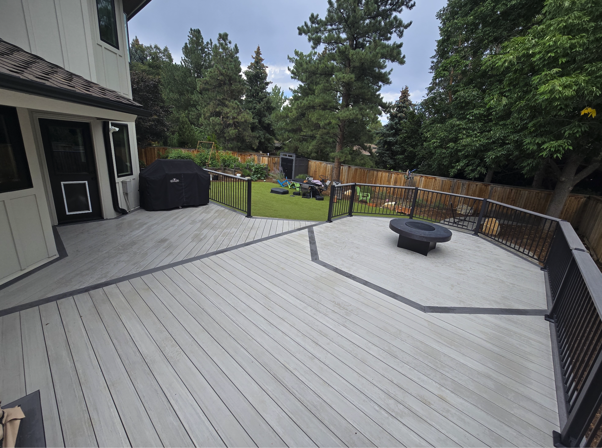 Large outdoor deck leading to a backyard with green grass, trees, a black grill cover, a firepit, and assorted outdoor furniture and toys.
