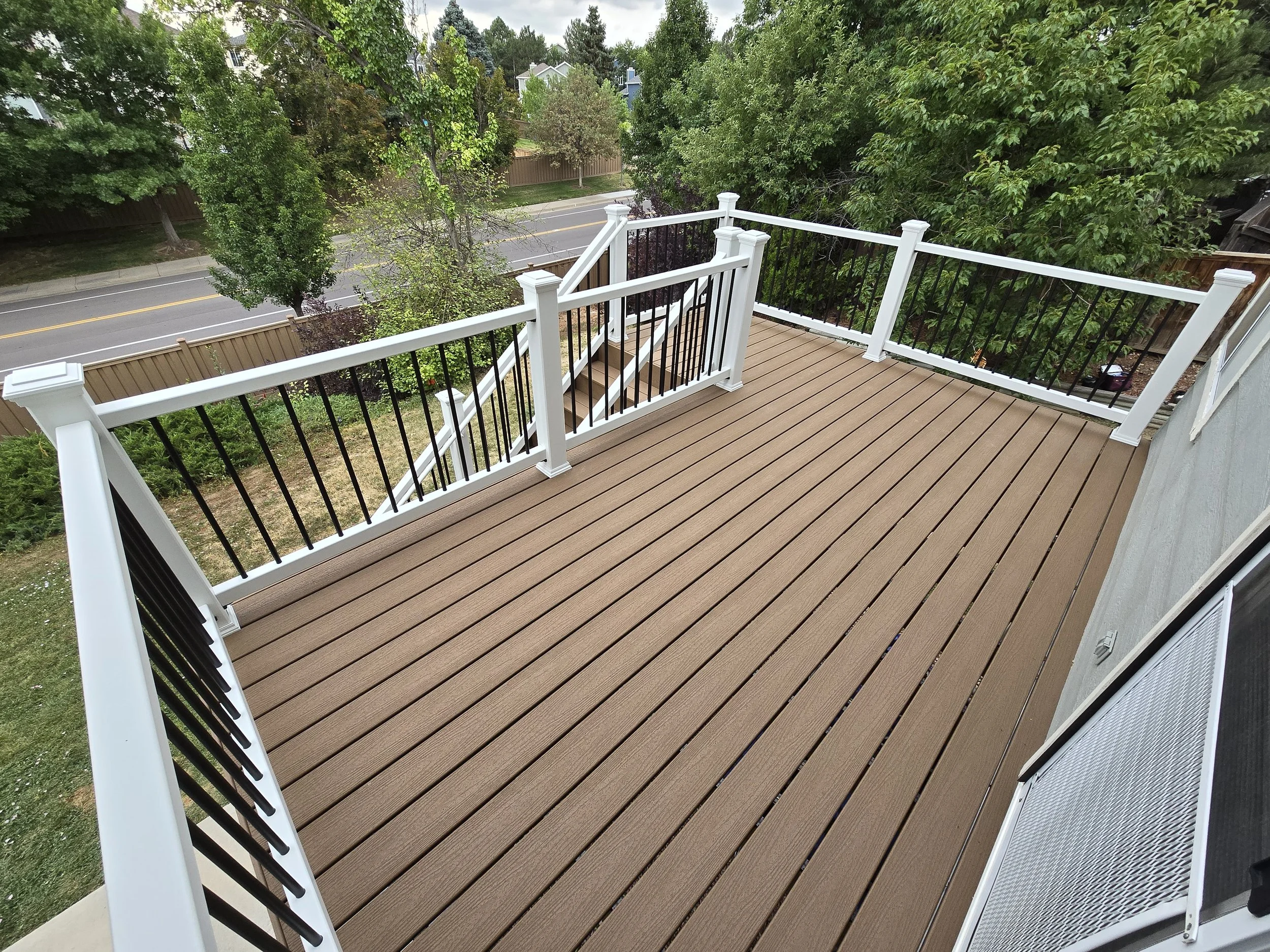 A spacious outdoor deck with white and black railing overlooking a suburban street and green trees. Trex Decking. Highlands Ranch