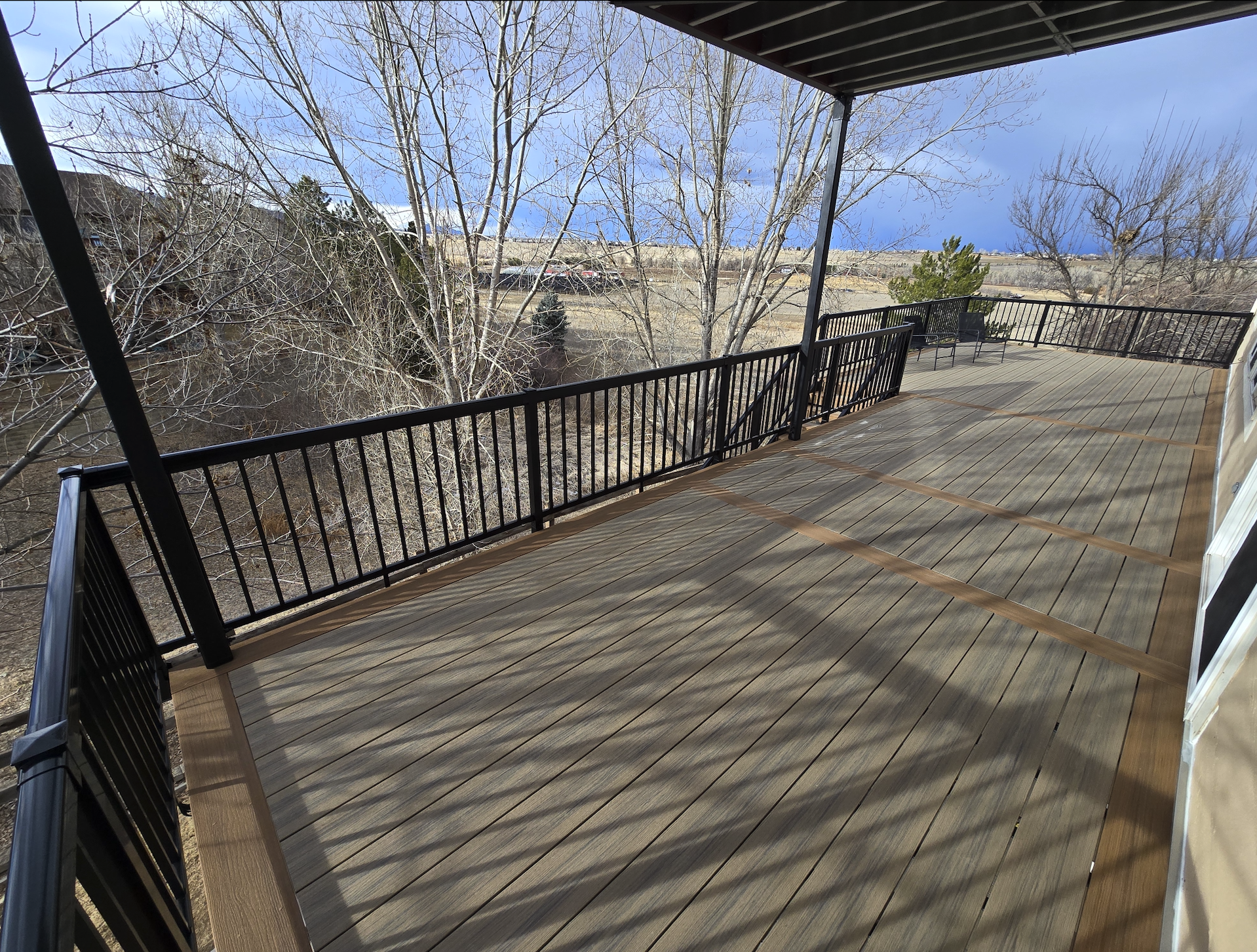 Outdoor deck with Deckorators decking and black metal railing, overlooking leafless trees and distant landscape under a partly cloudy sky. Littleton