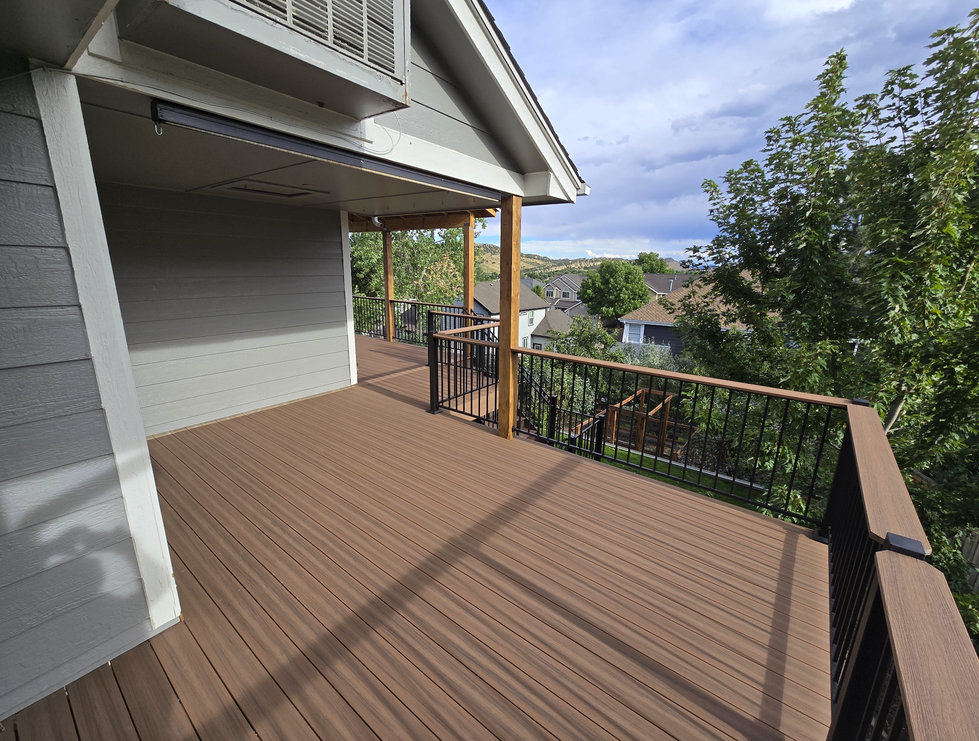 Empty wooden deck with black metal railings overlooking a residential neighborhood with trees and houses under cloudy sky.
