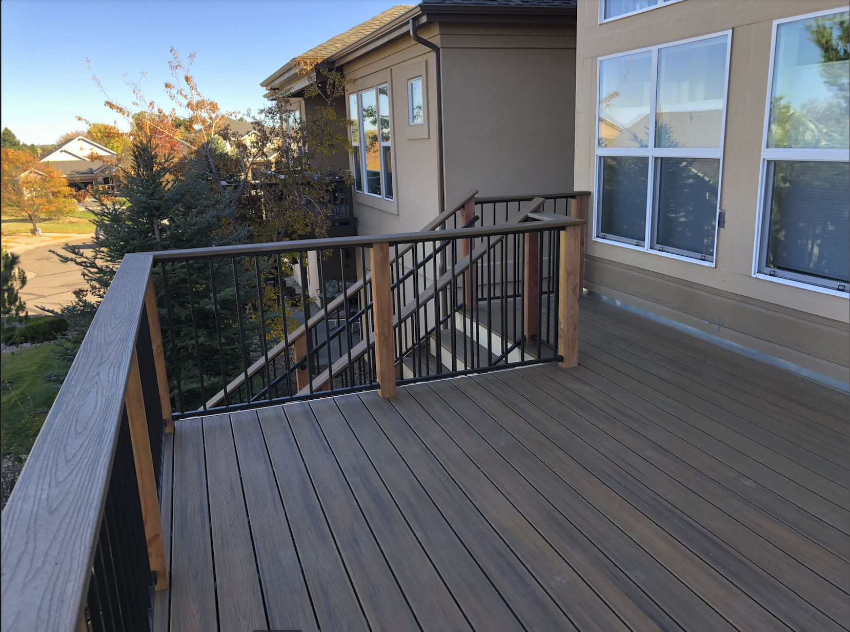 Newly built outdoor wooden deck with black metal railing, attached to a beige house with large windows, overlooking a neighborhood with trees and clear blue sky.