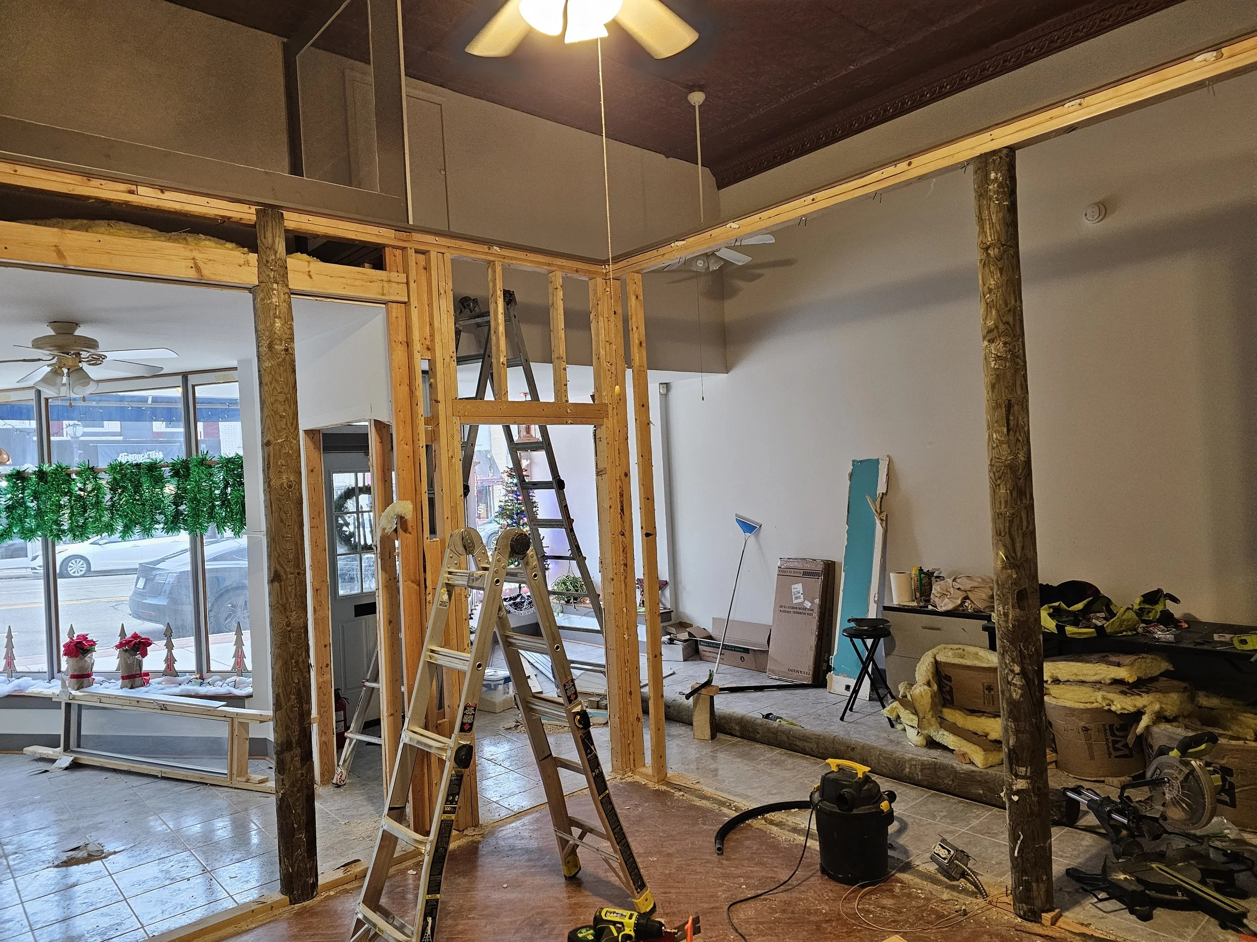 Interior of a building under renovation with exposed wooden framing, ladders, construction tools, and holiday decorations in the window.