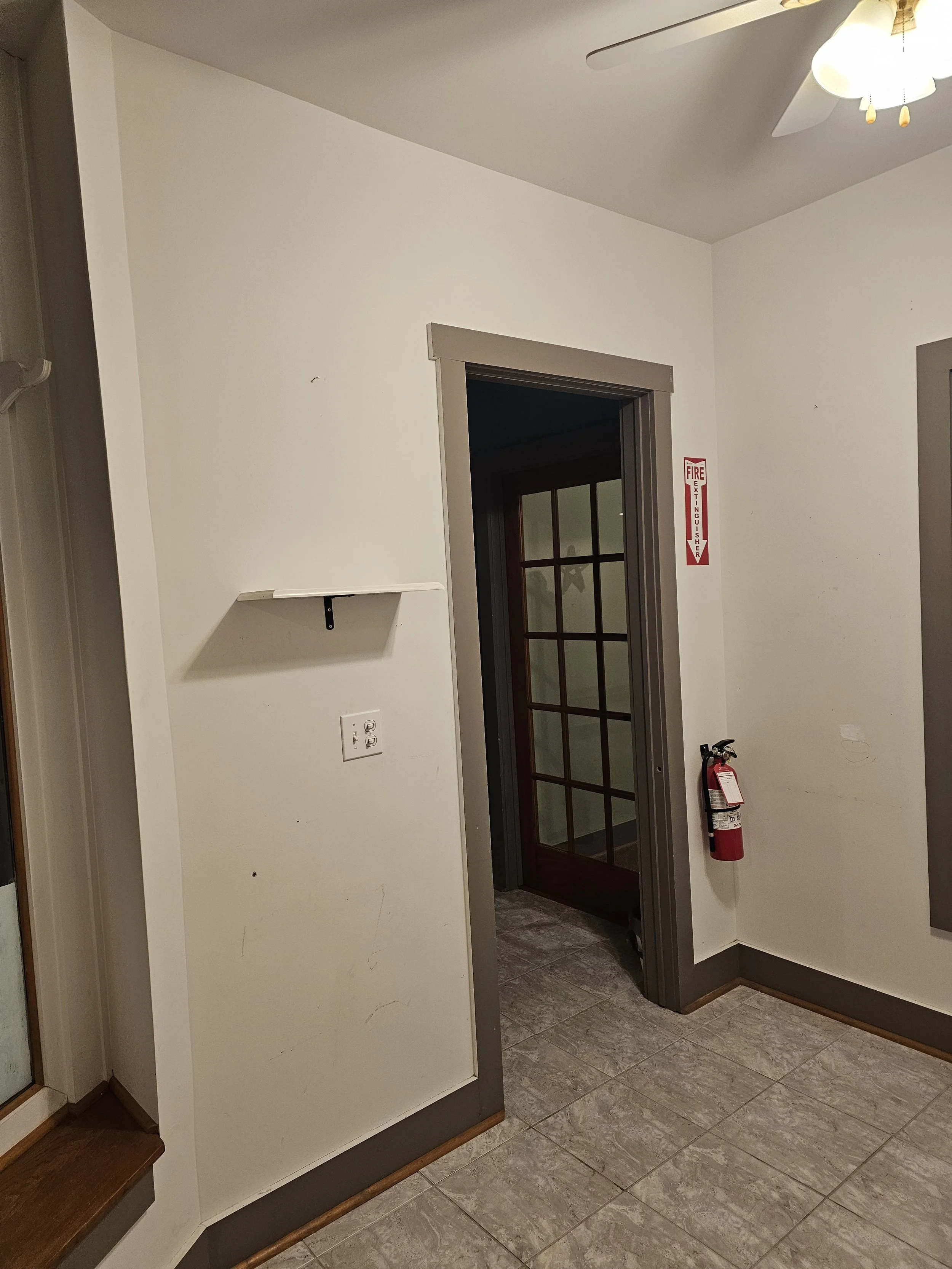 Interior of a room with beige tile flooring, white walls with gray trim, a fire extinguisher mounted on the wall, a ceiling fan with lights, and a doorway with a glass-paneled door leading to another room.