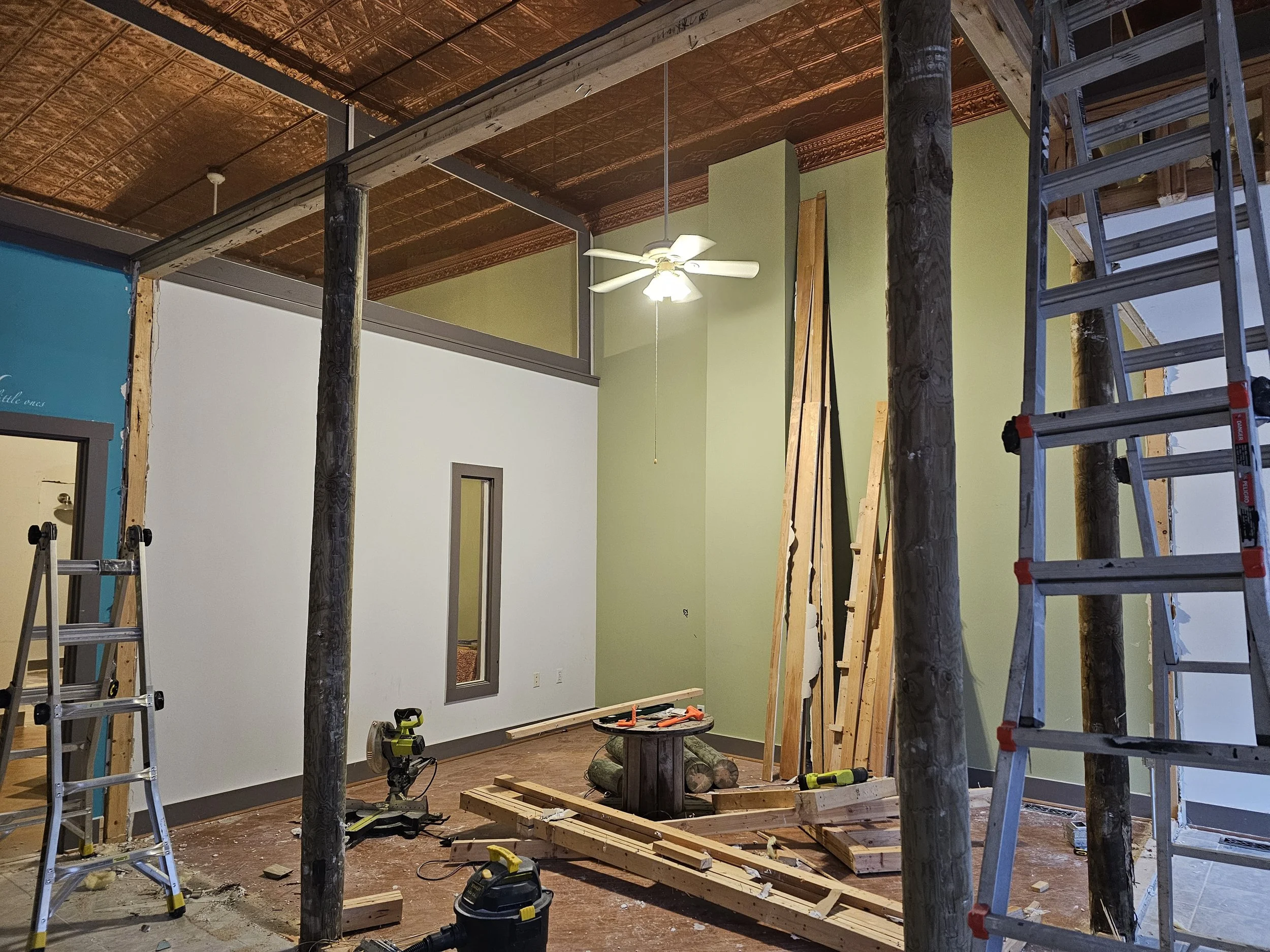 Interior of a room under construction with ladders, wood pieces, a ceiling fan, and tools.