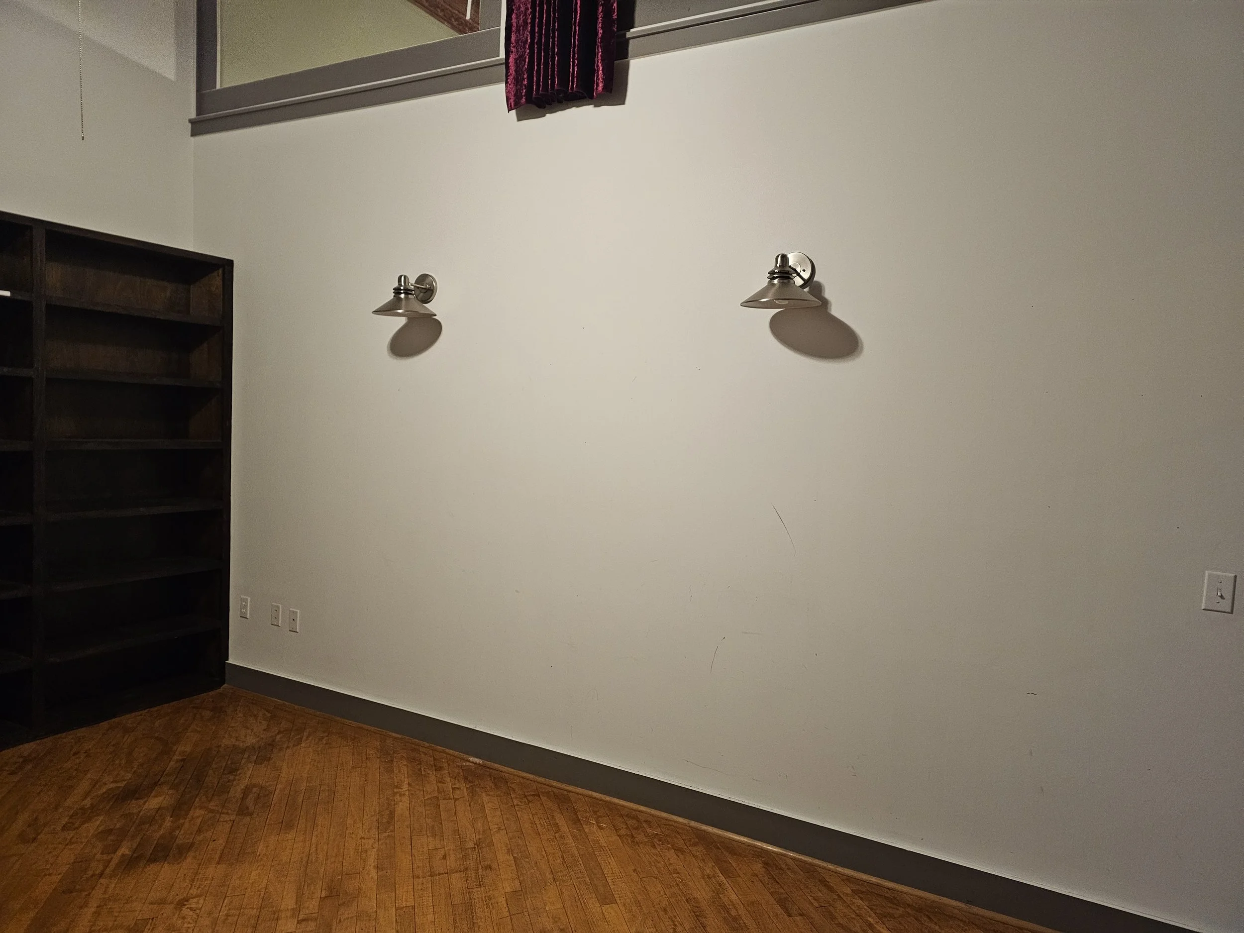 Empty room with two wall-mounted lights, a dark wooden bookshelf on the left, and a wooden floor.
