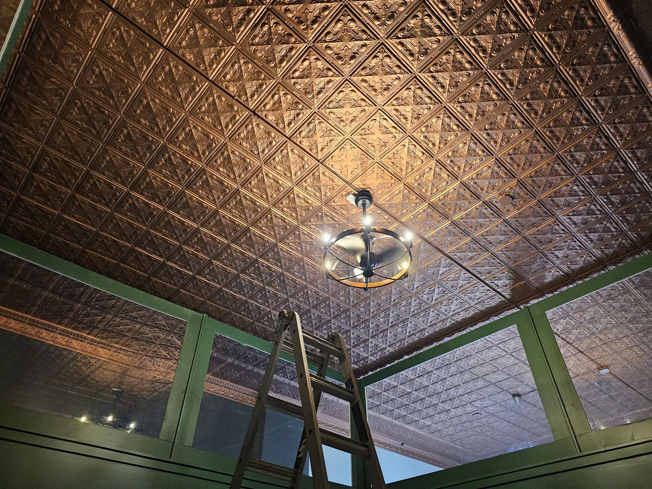 Interior view of a ceiling with decorative tin tiles and a ceiling fan with a light. A ladder is positioned in front of a large window with green trim.