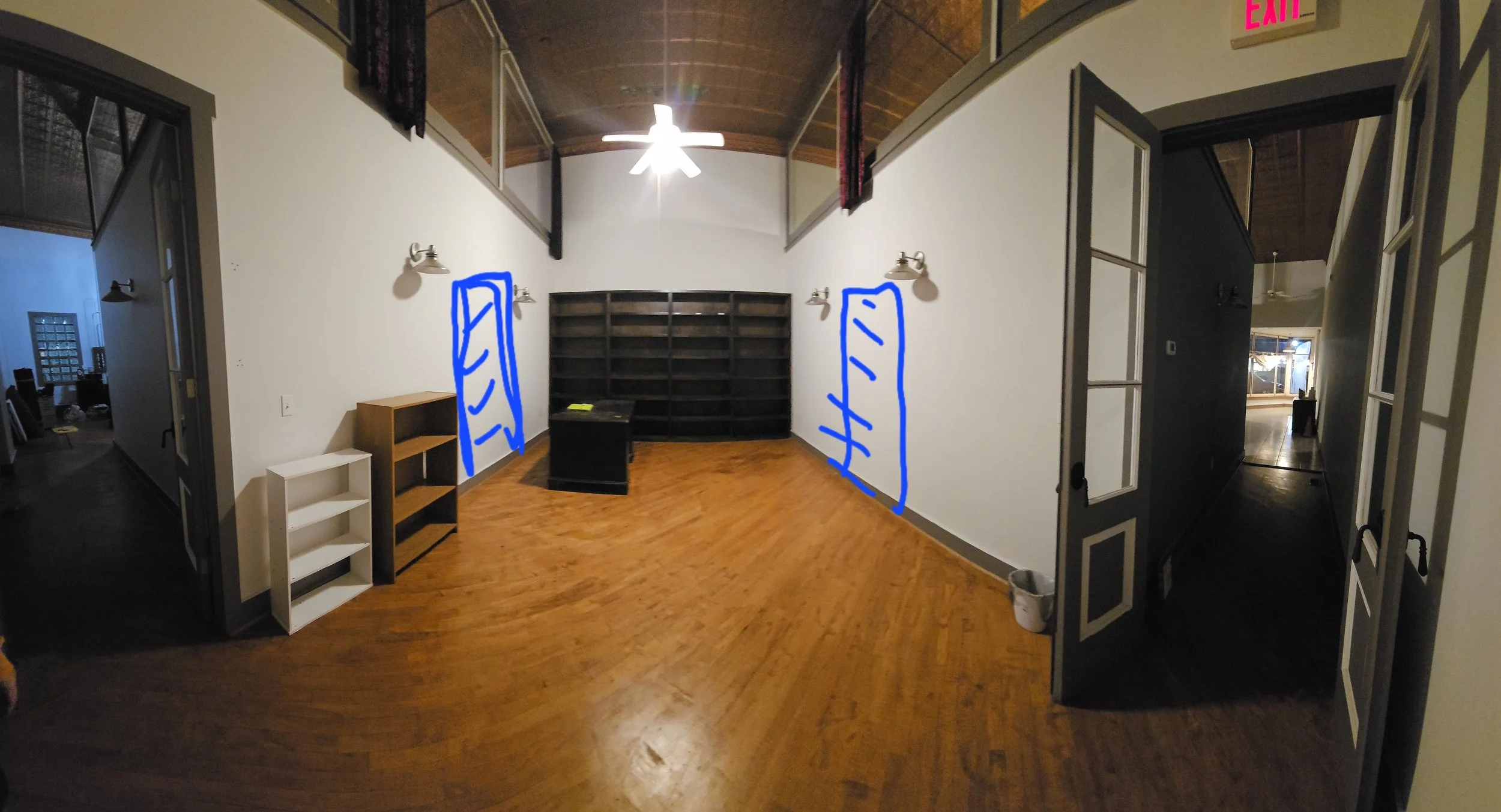 Empty room with hardwood floors, white walls, a large dark bookshelf, and two bookshelves marked in blue. There are wall-mounted lamps, a small desk, and an office chair. Hallways extend to the left and right, with the right hallway showing another r