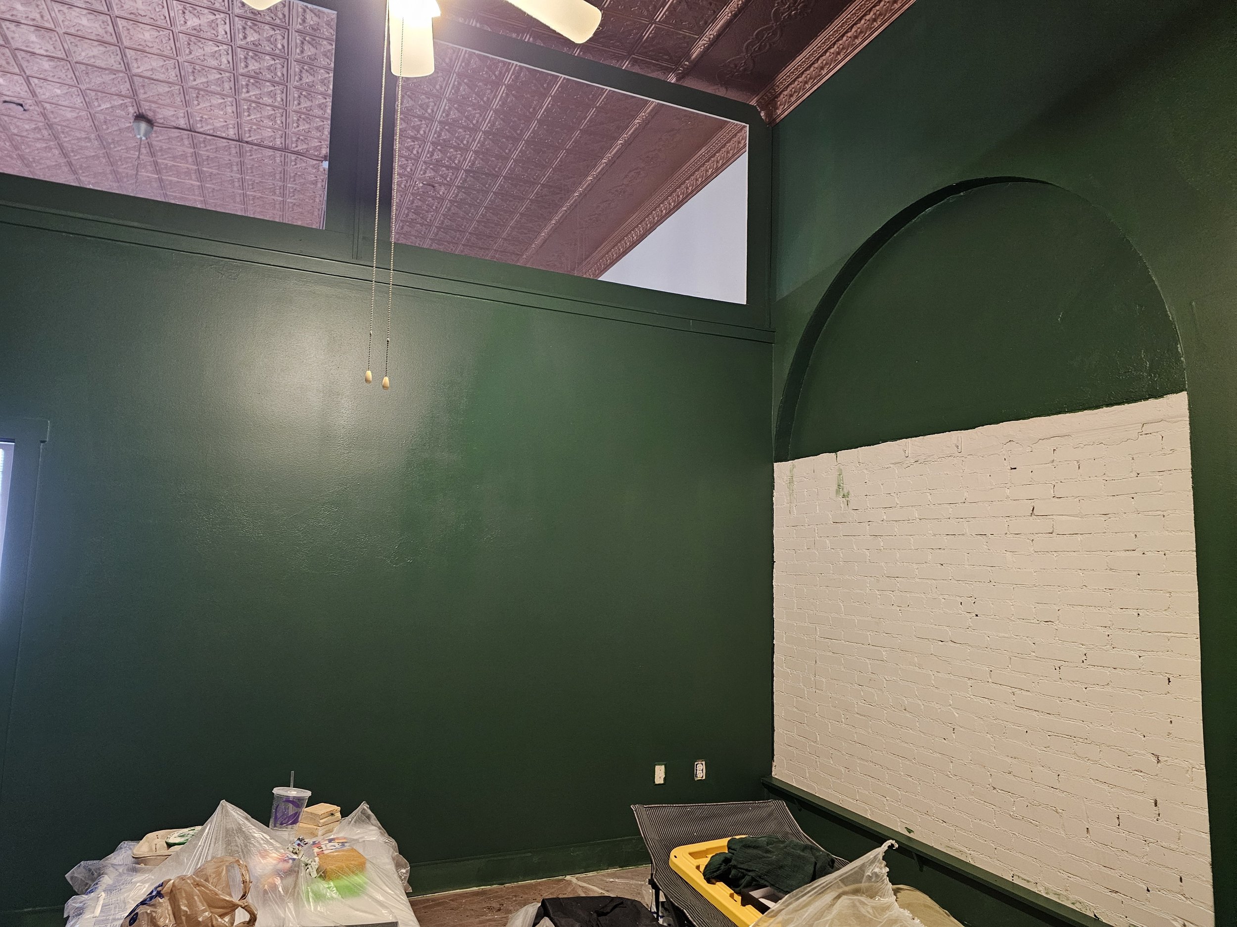 Interior of a room with dark green walls, a white brick accent wall, and a pink patterned ceiling with a ceiling fan. There are bags and tools on the floor, suggesting renovation or painting work in progress.