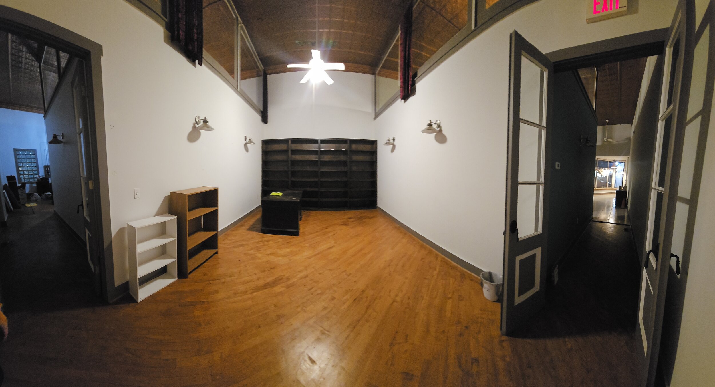 Empty room with wooden flooring, white walls, a black bookshelf, two small bookshelves, and a ceiling fan with attached lights. There are open doors leading to other rooms and hallways.