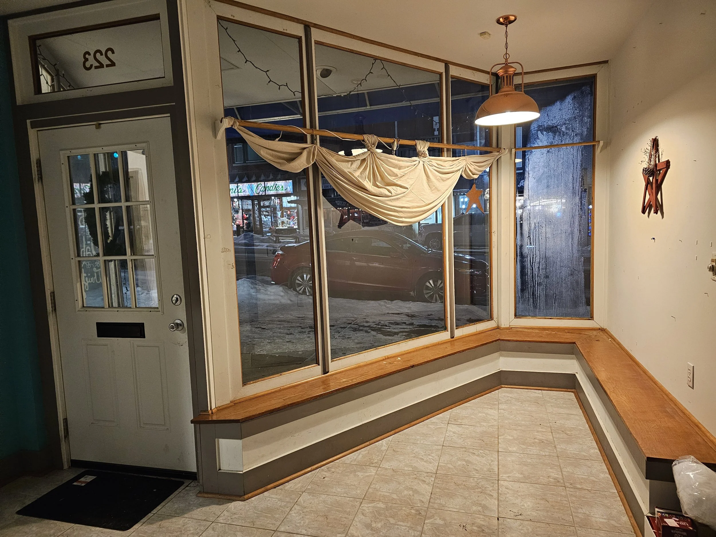 Empty interior corner of a small business with a large window, a hanging copper light, a white door with glass panels, and a built-in wooden bench along the walls. Outside, cars are visible through the frosted window, and there are snow and a store w