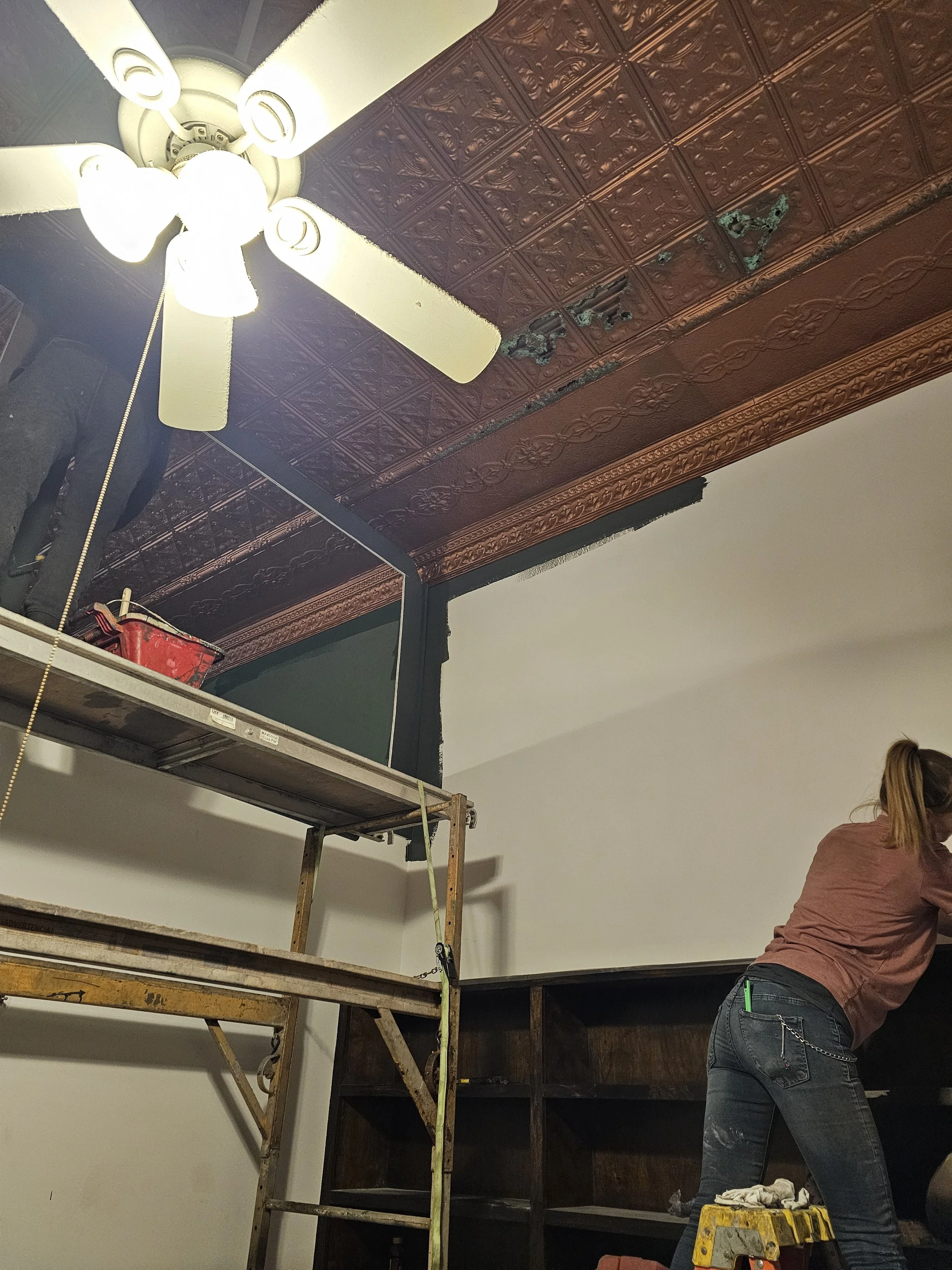 A woman works on renovation or maintenance inside a room. She is standing on a small yellow ladder, facing a dark wood cabinet. The ceiling has ornate, decorative tin tiles with some areas damaged or missing, revealing the structure underneath. A cei