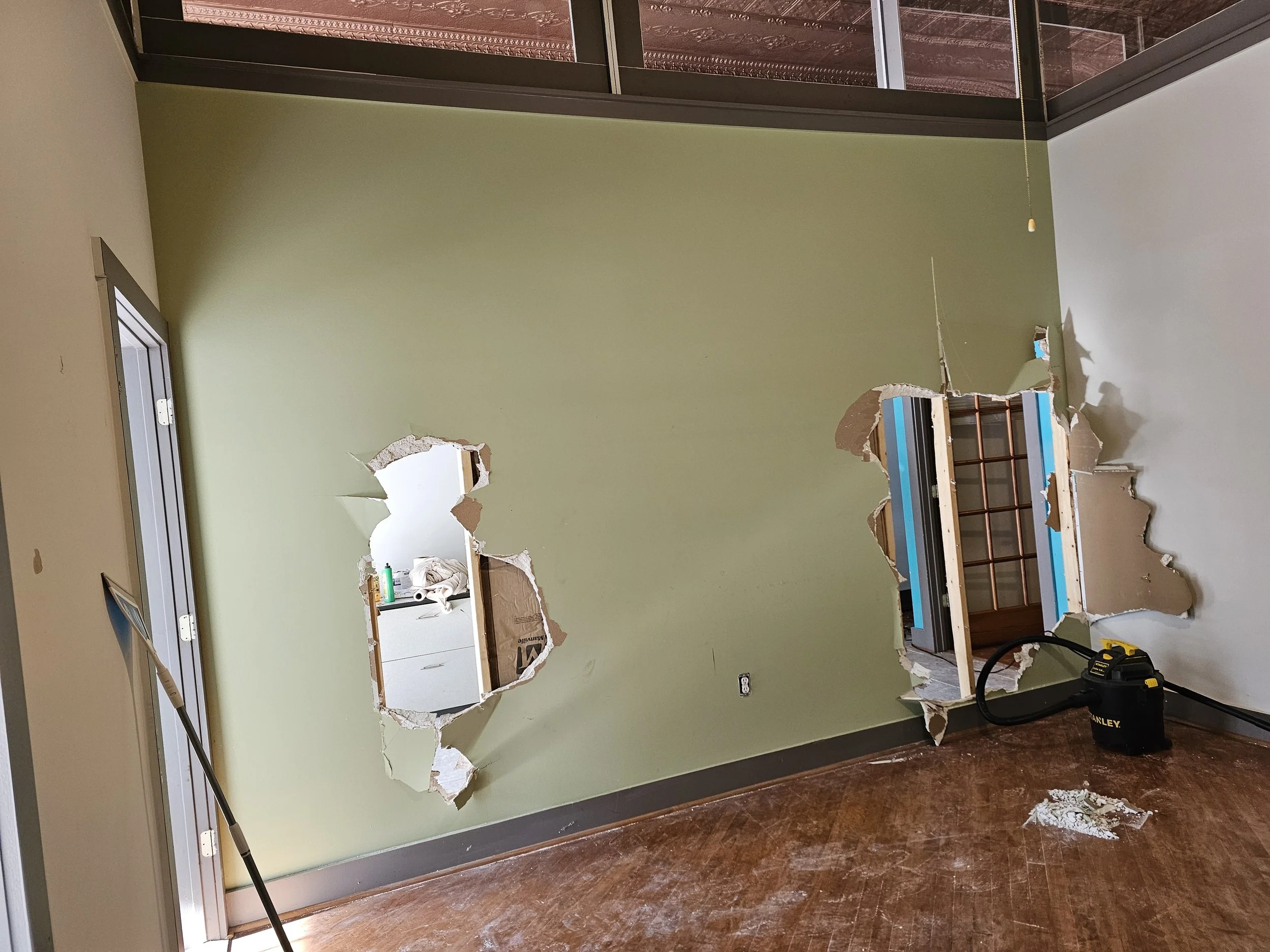 Room with two large holes in the wall, exposing interior spaces with visible framing and utilities. The room has green and white walls, a wooden floor, and a window or door frame on the left side. Construction tools and debris are present, indicating