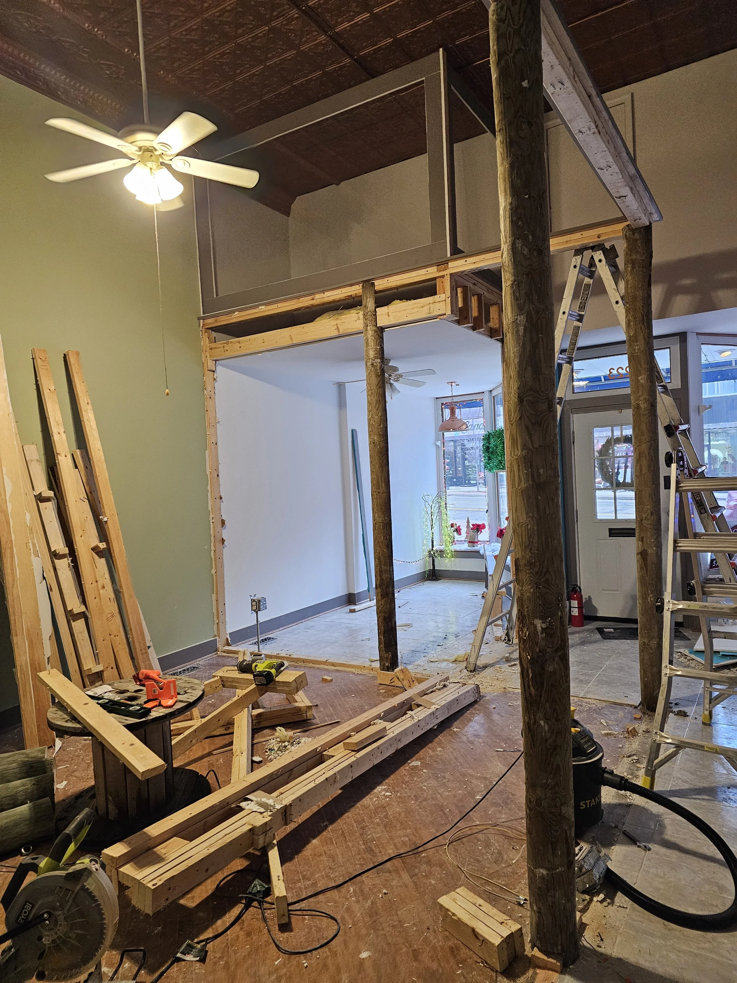 Interior view of a building under renovation with exposed wooden beams, tools, a ladder, and construction materials. There is a partially framed wall, a ceiling fan, and a window with plants outside.