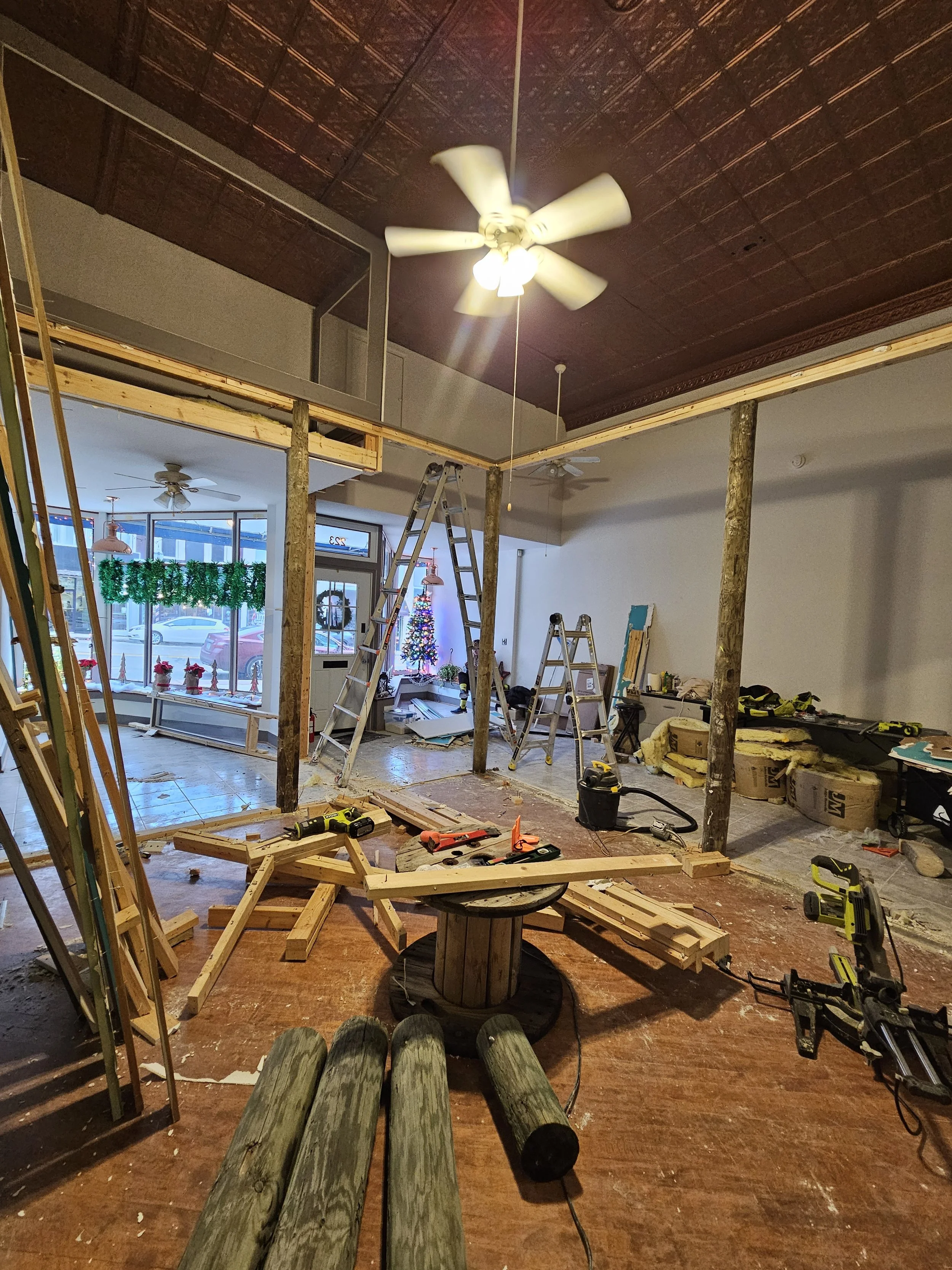 Interior of a building under renovation, with wooden beams, ladders, tools, construction materials, and a small Christmas tree near the entrance.