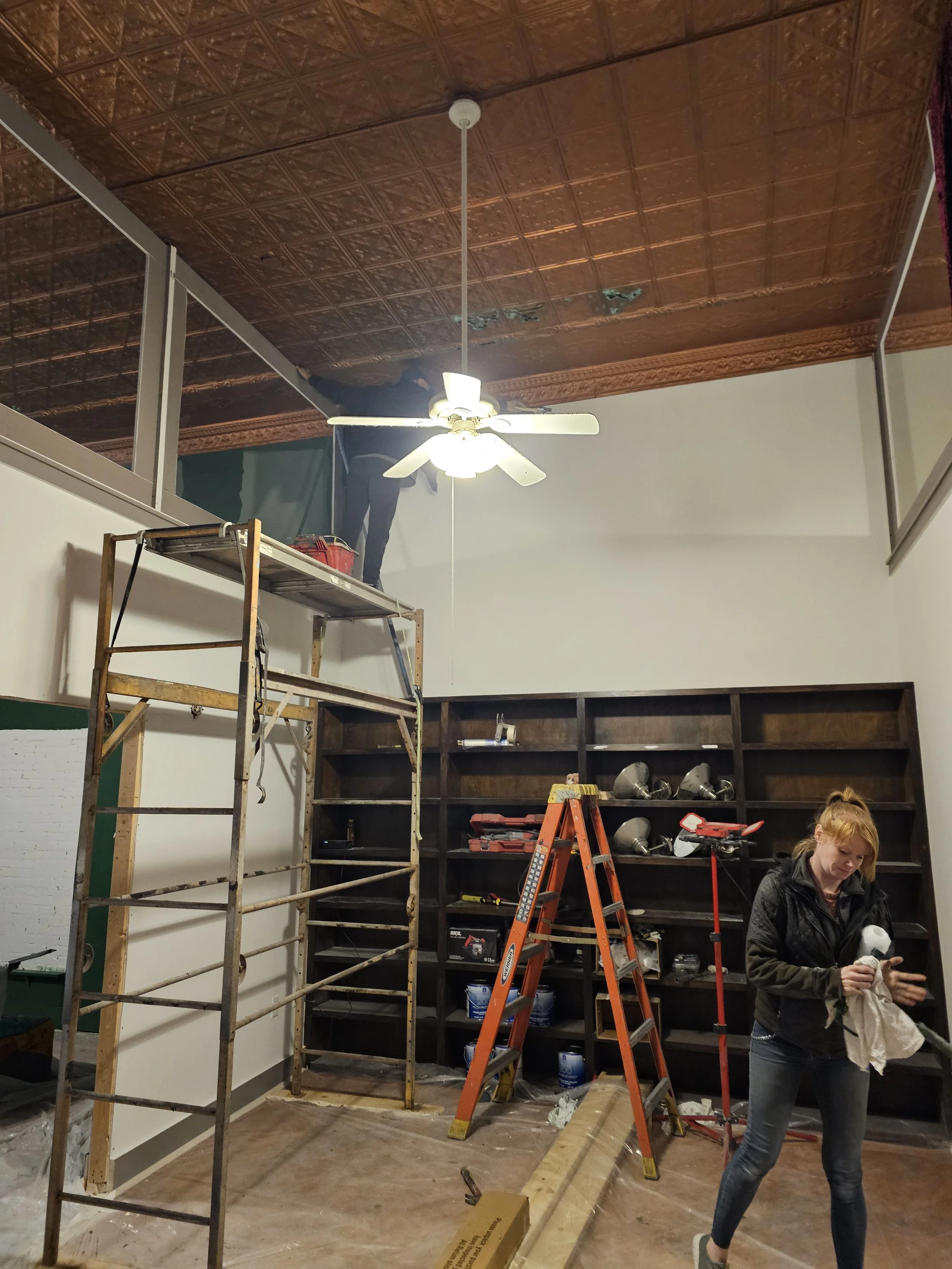 Woman standing in a room under renovation, holding a paint roller and cloth, with construction tools and ladders around her.