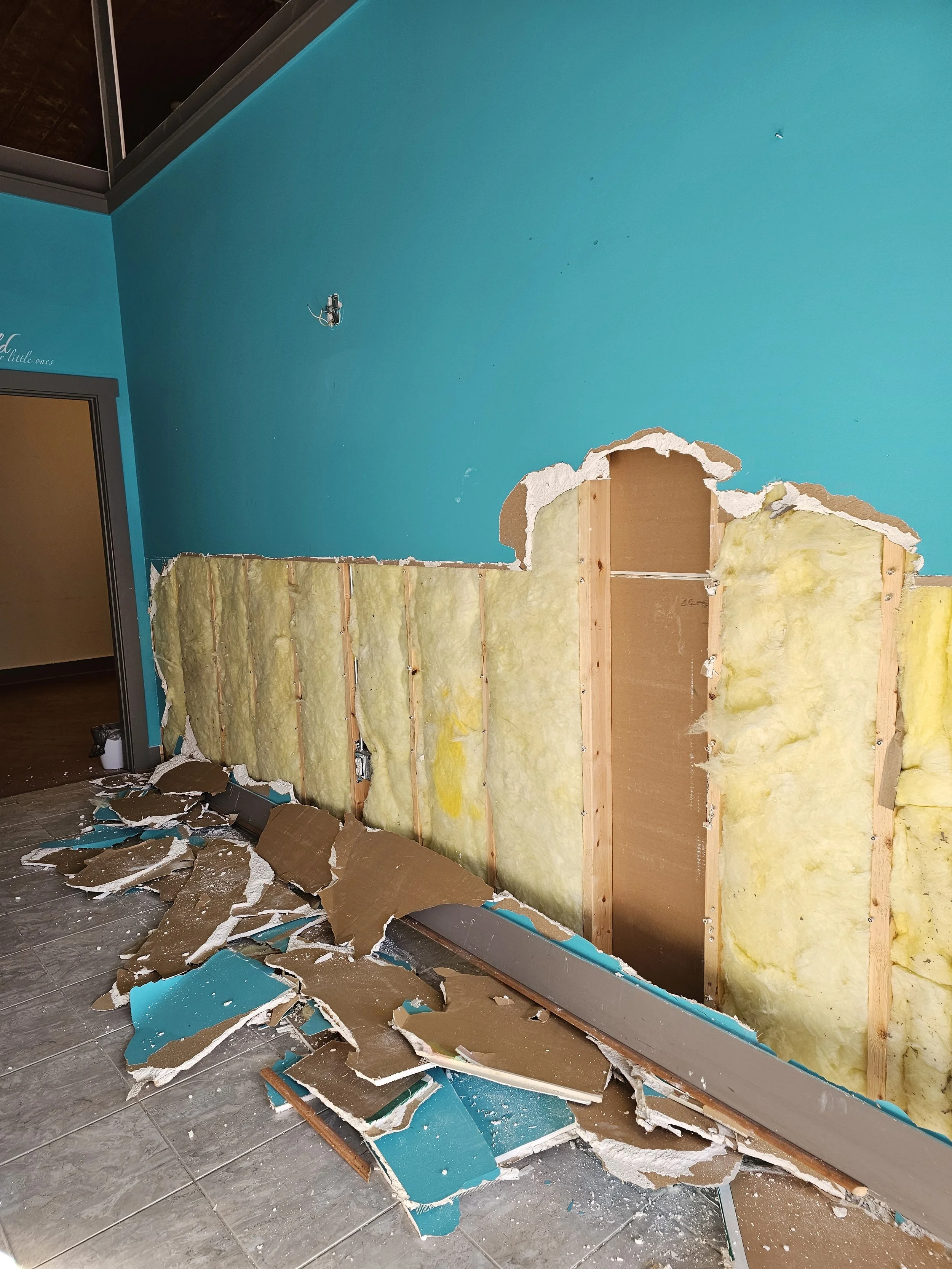 A partially demolished wall with exposed insulation and wooden studs in an indoor space with blue painted walls, debris on the floor, and an unfinished or damaged section.