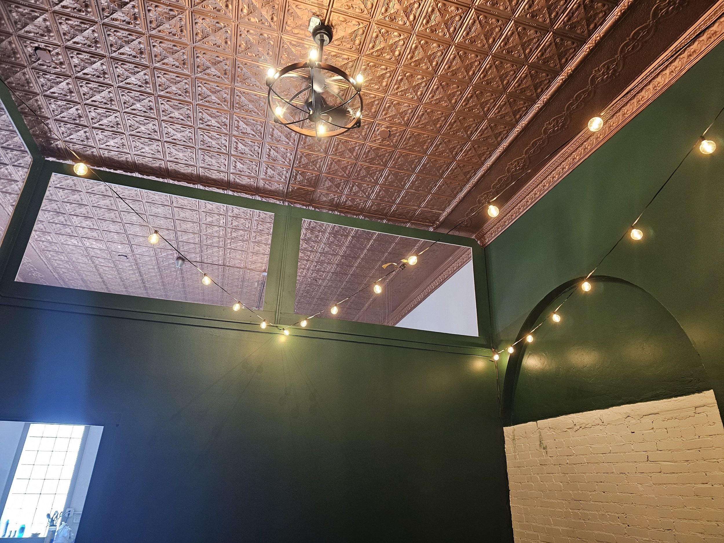 Interior ceiling with decorative tin tiles, green walls, exposed brick, string lights, and a black ceiling fan with light fixtures.