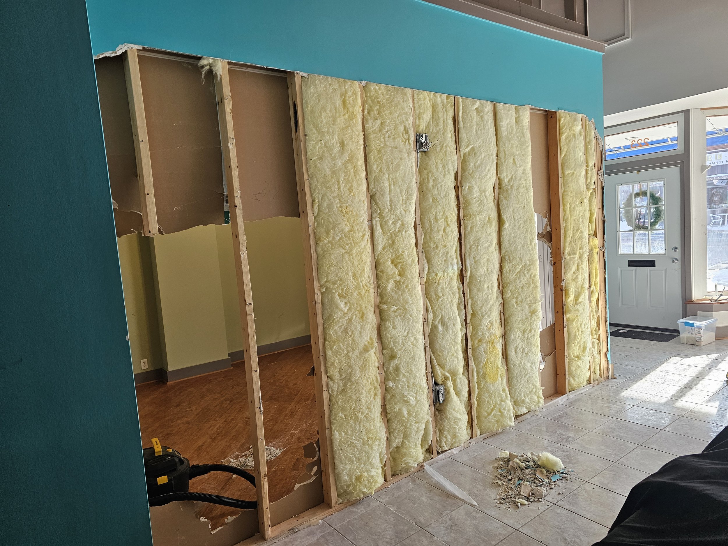 Interior wall with insulation being installed, exposed studs and electrical outlets, in a room with a tiled floor and a glass door entrance.