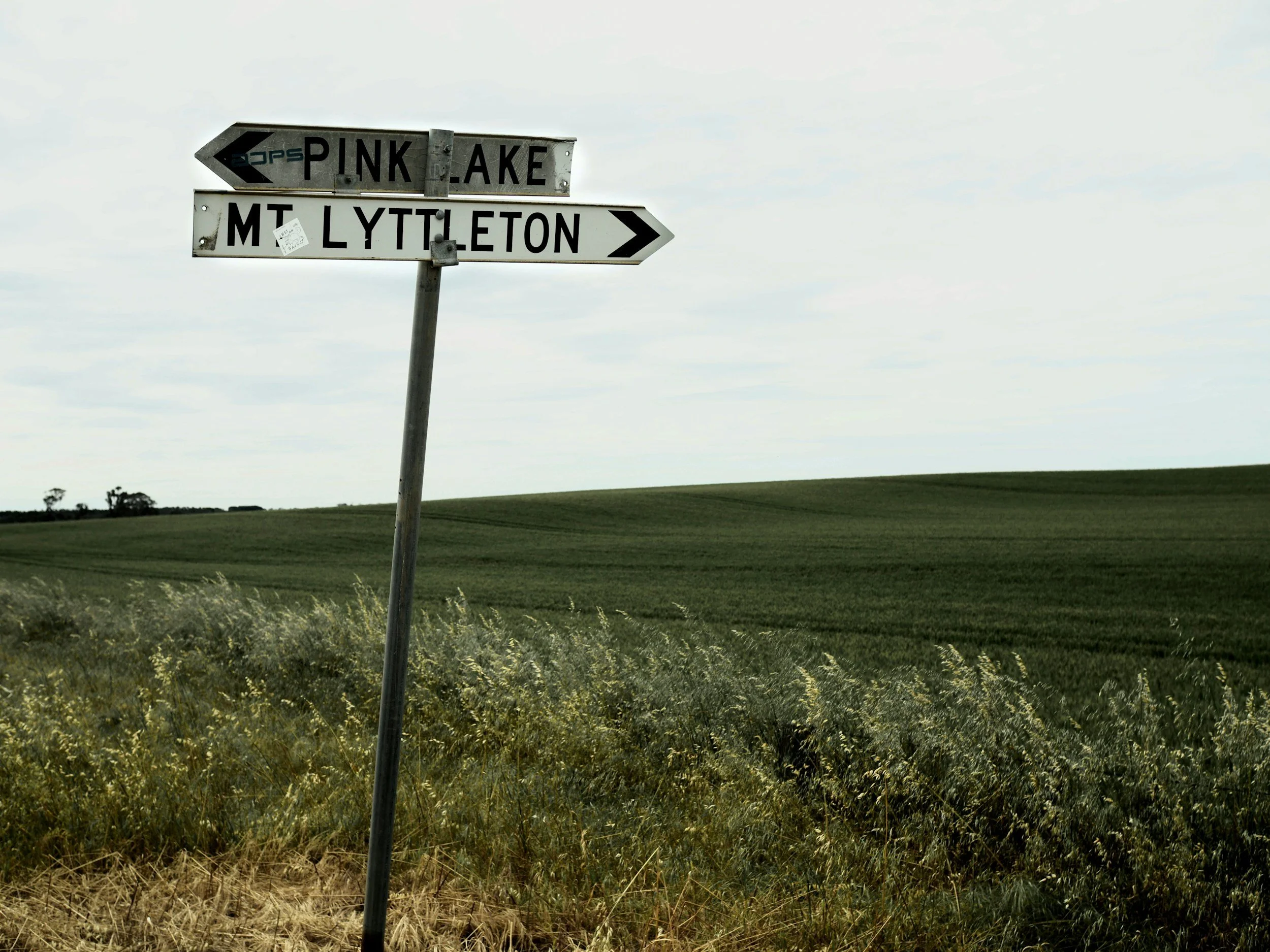 A street sign with directional arrows pointing towards Pink Lake and Mt. Lytleton, set against a backdrop of green fields and a cloudy sky.