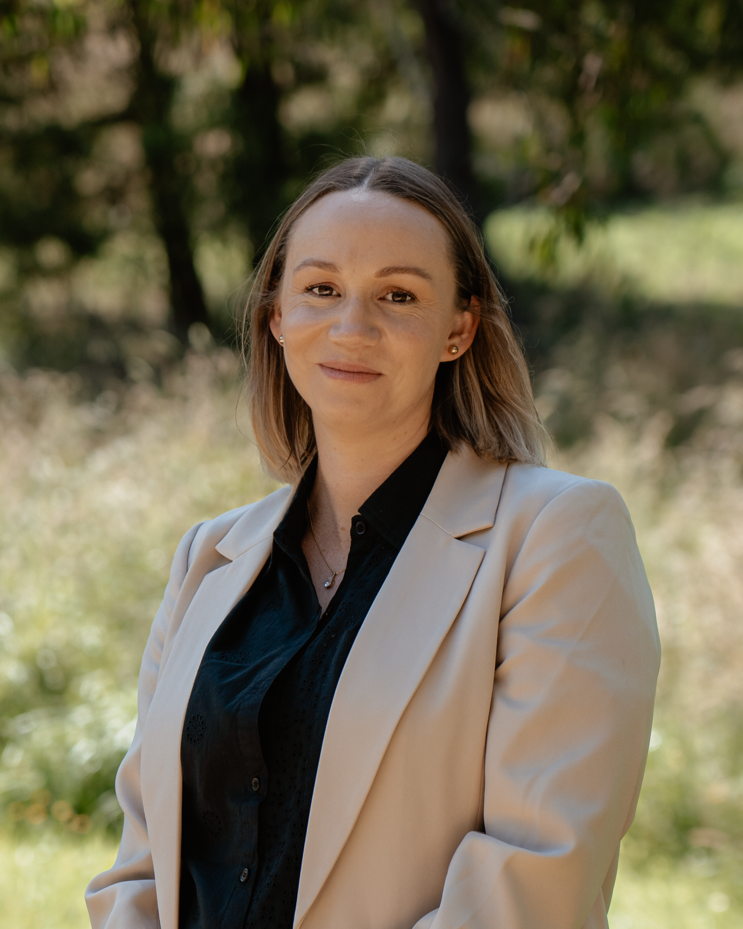 Yarnie Couchman with shoulder-length blonde hair, wearing a beige blazer over a black blouse, standing outdoors in a park with trees and greenery in the background. She has a subtle smile and is looking at the camera.