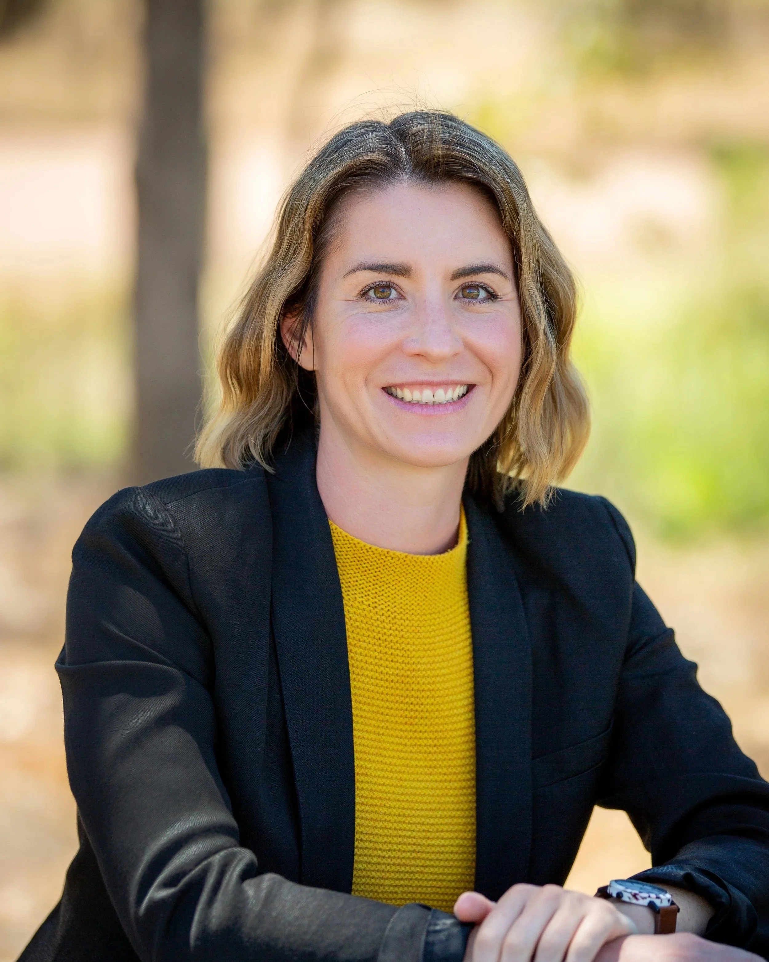 Pru Cook with shoulder-length wavy hair, wearing a black blazer and a yellow top, smiling outdoors with a blurred background of trees and leaves.