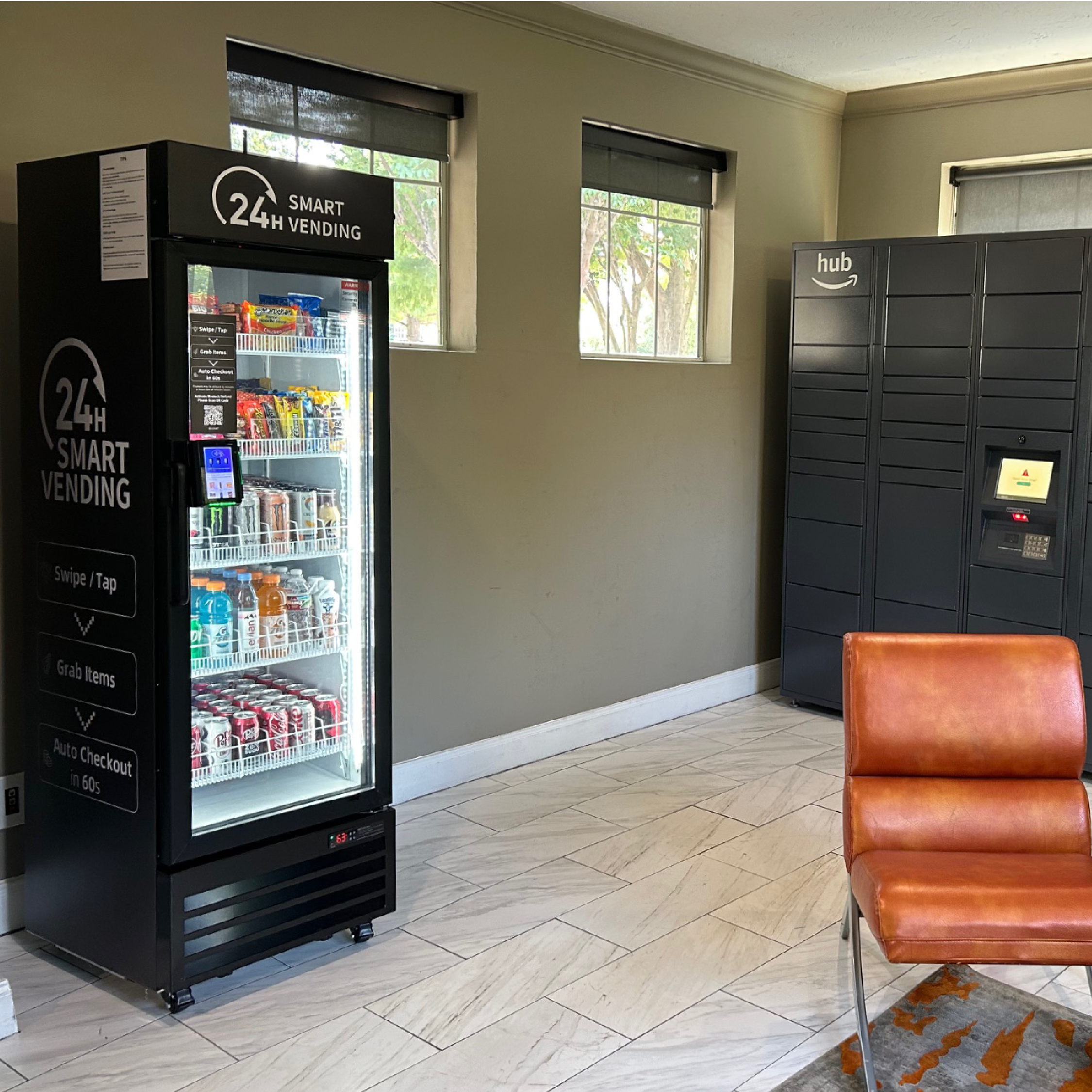 Interior of a vending area featuring a black 24-hour smart vending machine stocked with snacks and drinks, a dark gray Amazon hub locker, and an orange leather chair on a patterned rug, with two small windows on a beige wall.