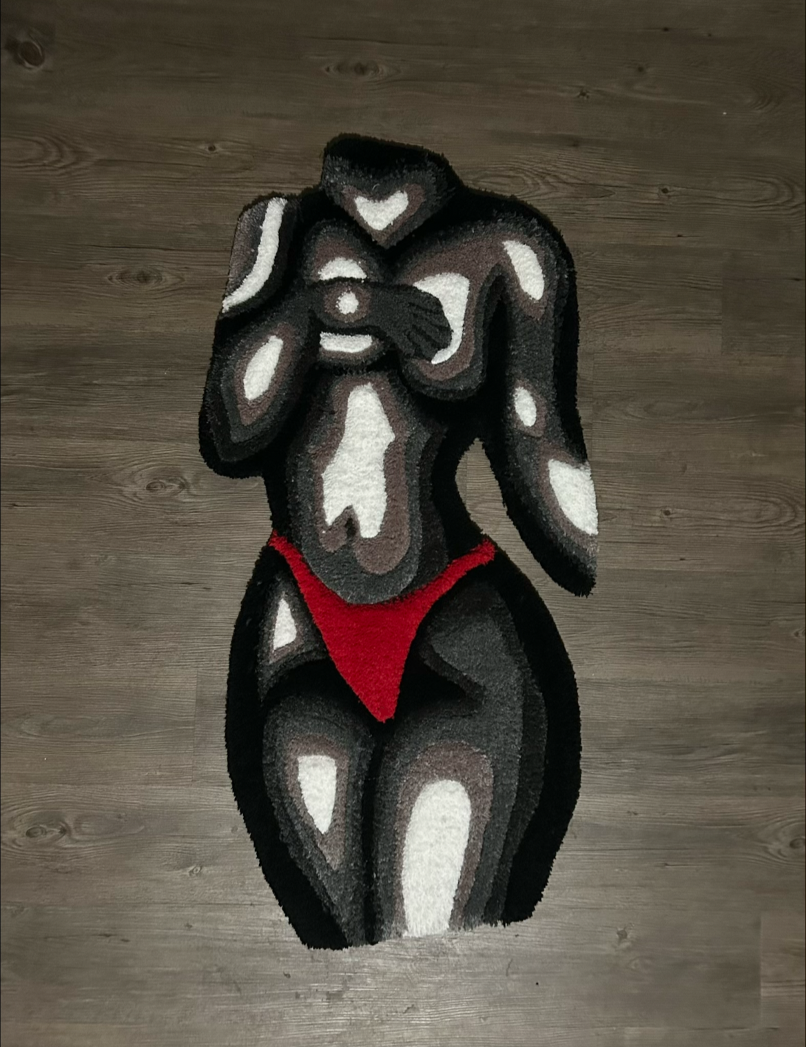 A rug designed like a stylized, abstract female figure with black, brown, white, and red colors, covering her face with her hand, wearing red underwear, on a wooden floor.