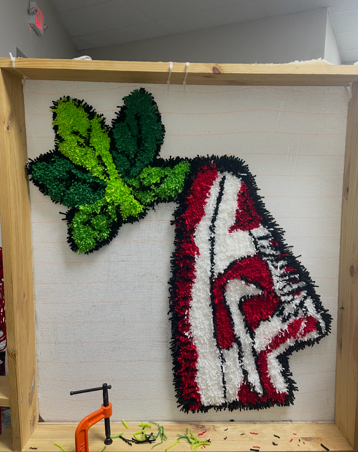 A large decorative piece made of colorful materials depicting a green butterfly next to a red and white striped mitten, mounted on a neutral background within a wooden frame.