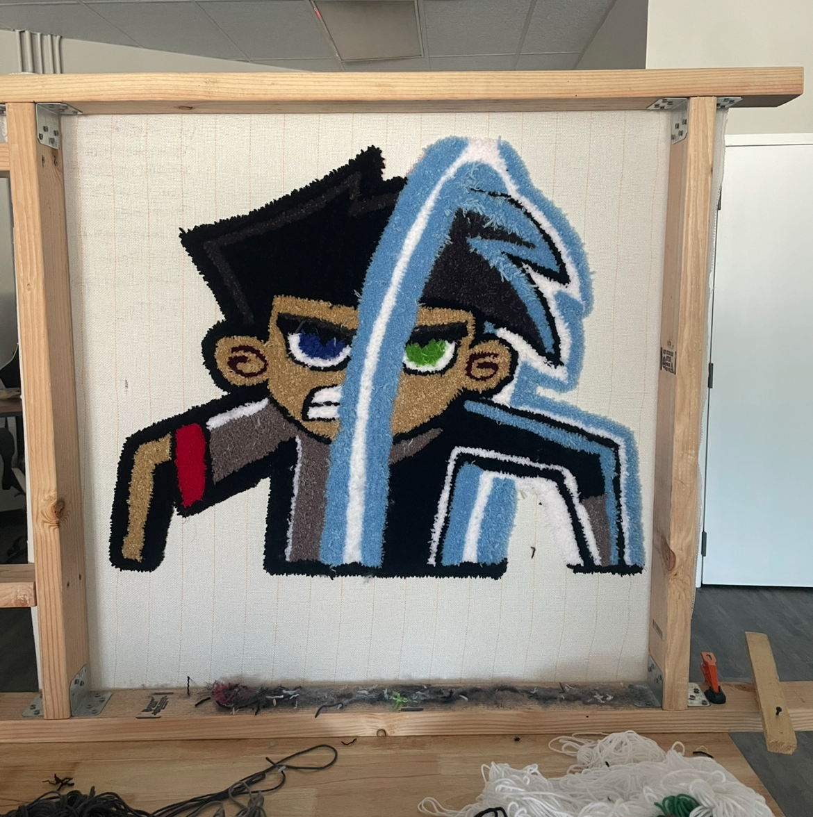 A pixel art-style embroidery of a character with black hair, an angry expression, green and blue eyes, wearing a brown shirt with a red strap, and holding a sword and shield, mounted on a wooden frame.