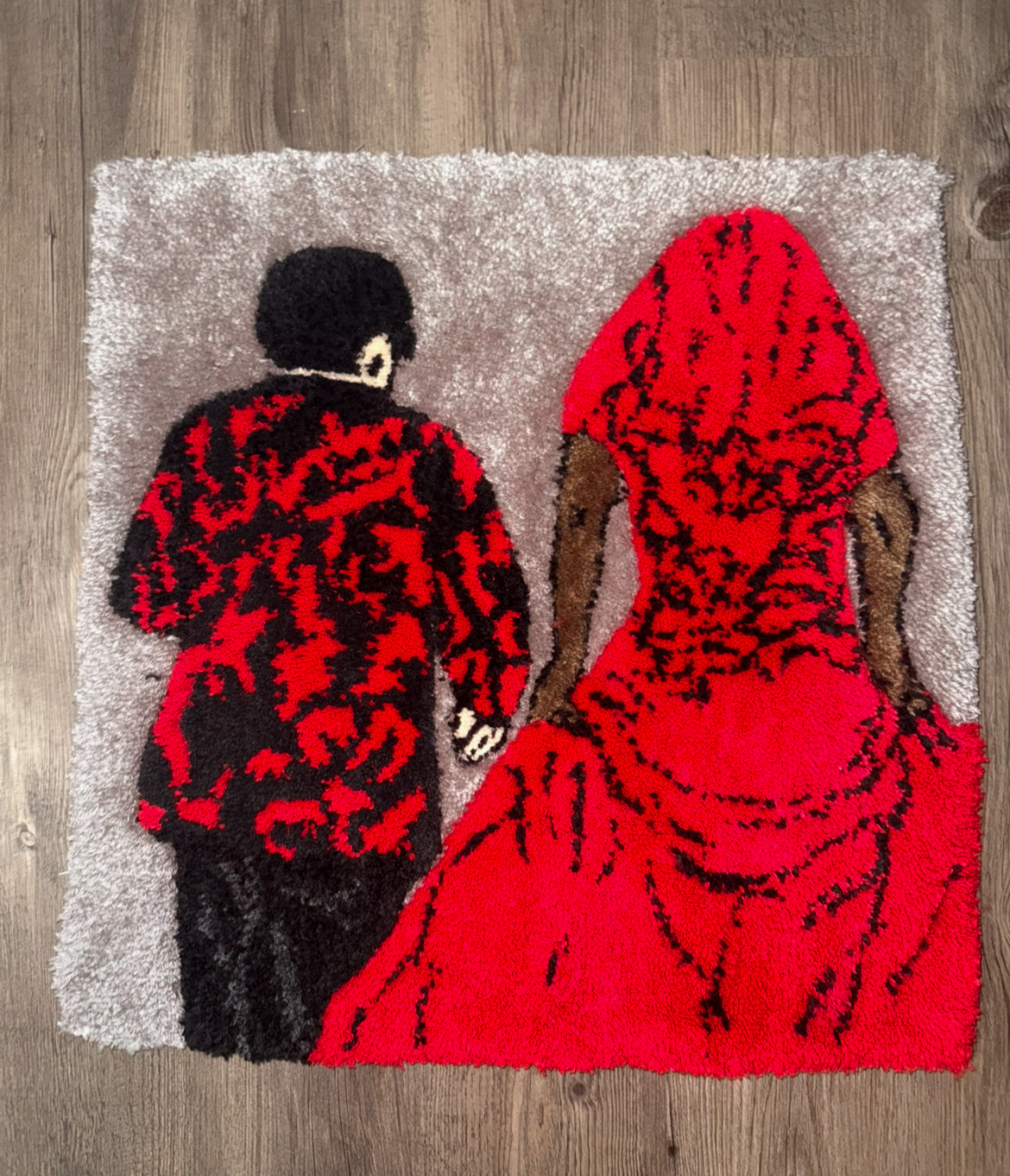 Rug depicting a man and woman holding hands, seen from behind. The man is in a red and black patterned jacket, and the woman is in a red dress with a hood, placed on a light-colored textured rug on a wooden floor.