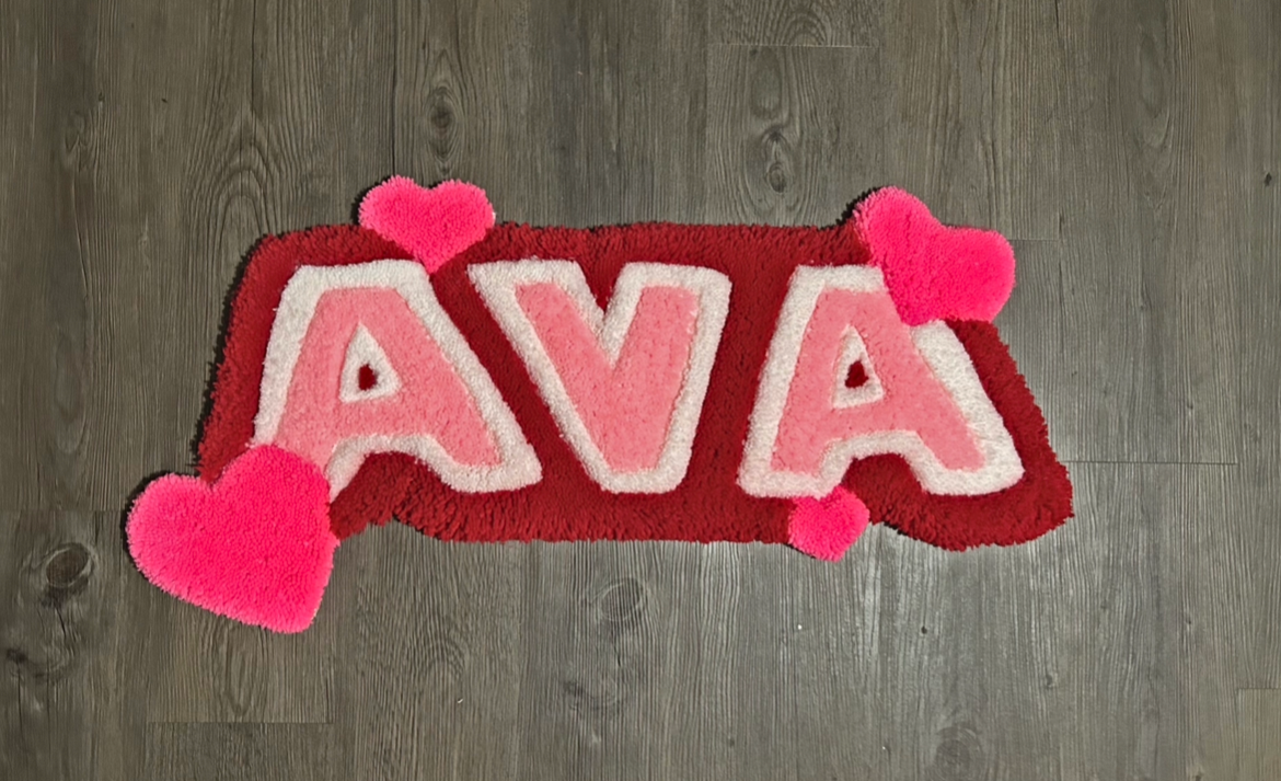 Rug with the name AVA in pink and white letters, surrounded by red background and pink heart shapes, on a wooden floor.