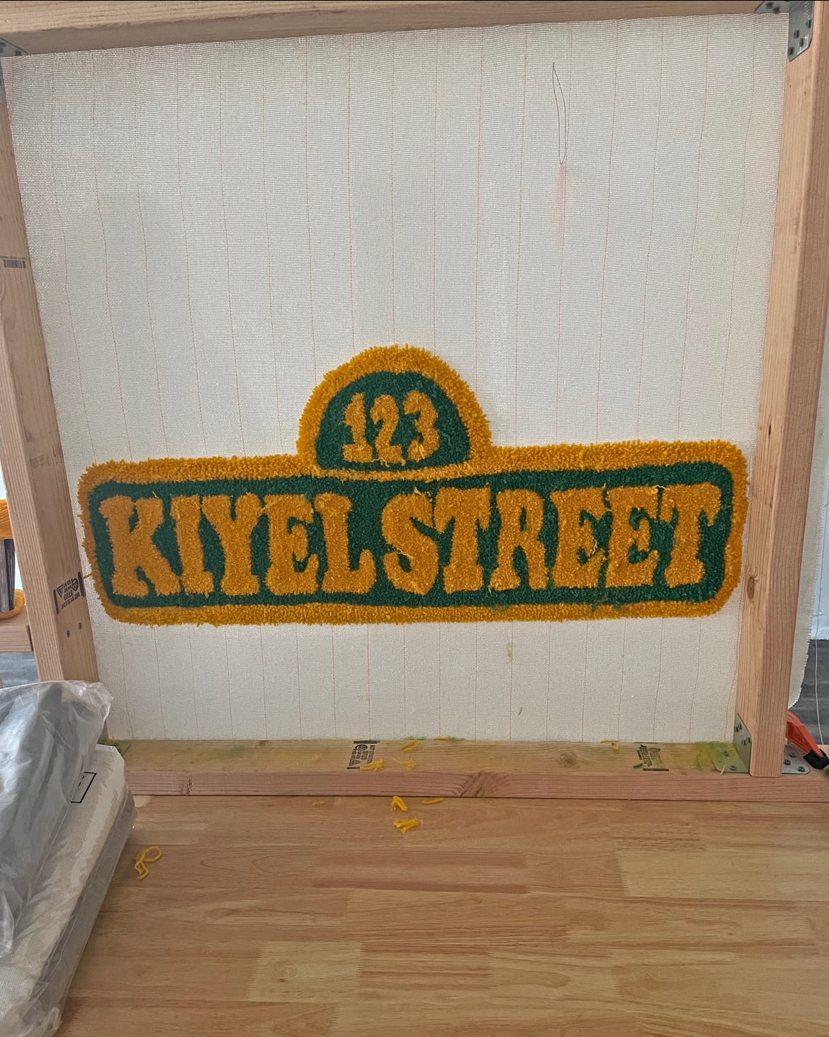 A decorative Welcome mat with the text "123 KIEL STREET" in orange and green colors, placed on an unfinished wooden wall.