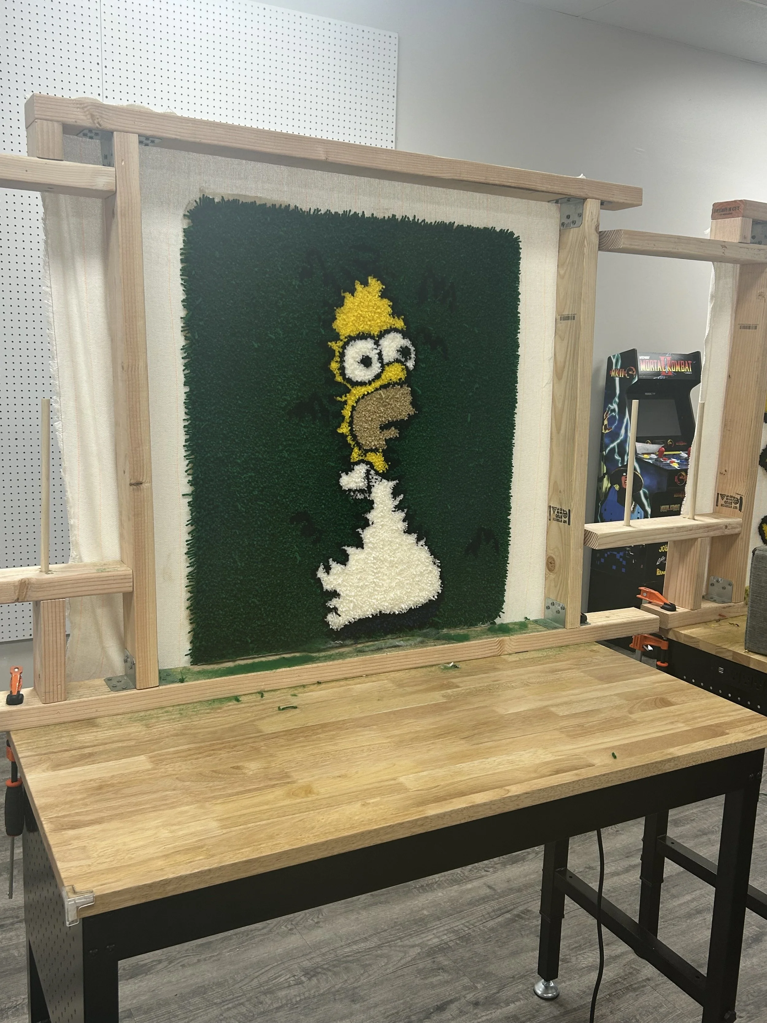 A pixel art of Homer Simpson made from colored materials, positioned on a cloth panel supported by a wooden frame, with an arcade game in the background.
