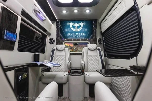 Interior view of a luxury van with beige diamond-stitched seats, fold-out tables, a large screen displaying 'Ultimate Toys,' and various entertainment features.