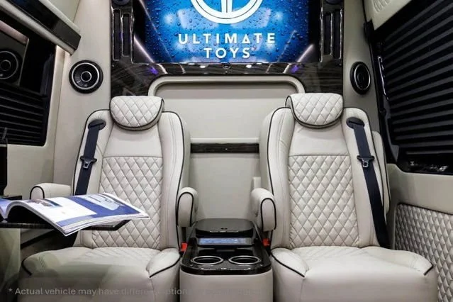 Luxury trailer interior with two beige, quilted leather seats with headrests, a small black table between the seats, and a large screen with the Ultimate Toys logo on the wall.