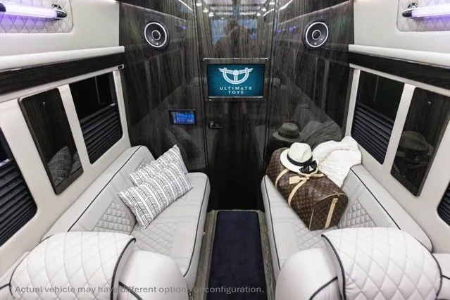 Interior of a luxury vehicle with two white leather seats, pillows, a brown bag, hats, a screen on the back wall, and black window shades.