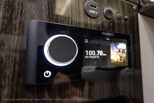 A Fusion audio control panel with a large circular volume knob, a small display screen showing radio station 100.7 FM, to the right of the knob.