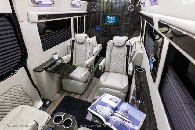 Interior of a luxury van with two beige quilted seats, a foldable tray table, a book, and a small cup.