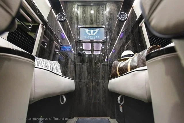 Interior of a luxury van or bus, showing two beige leather seats facing each other, with a wood-paneled wall at the back that has a screen displaying the Ultimate Journeys logo, and air conditioning vents.