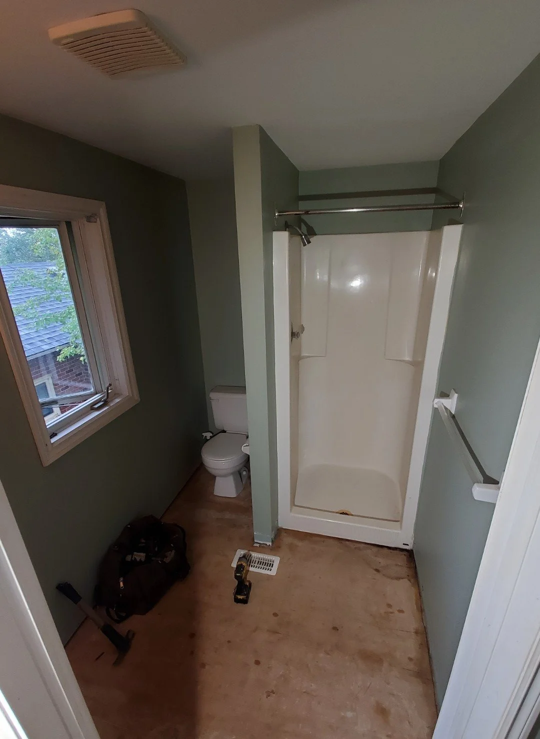 Bathroom with green walls, window, toilet, and shower stall. Floor is unfinished wood, tools and a backpack are present.