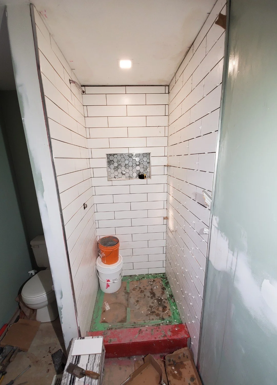 A bathroom under renovation with white subway tiles installed on three walls, a small niche with hexagon tiles, exposed drywall, construction tools, buckets, and debris on the floor.